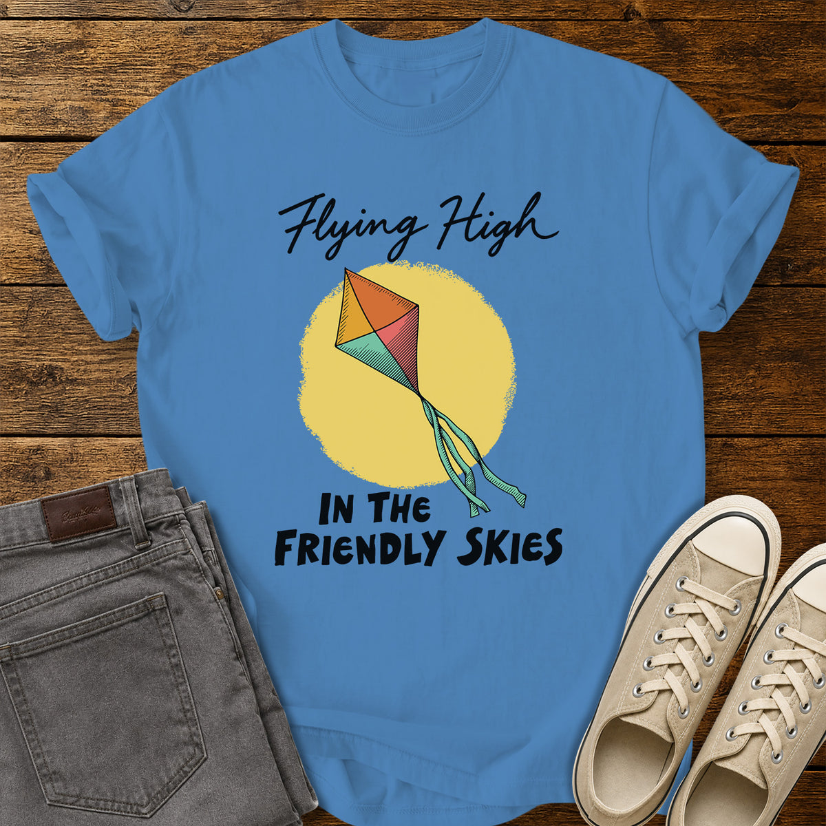 royal caribe blue T-shirt featuring a colorful retro kite flying against a yellow sun, with black script and bold lettering that reads “Flying High in the Friendly Skies,” displayed on a rustic wooden background with gray jeans and beige sneakers.