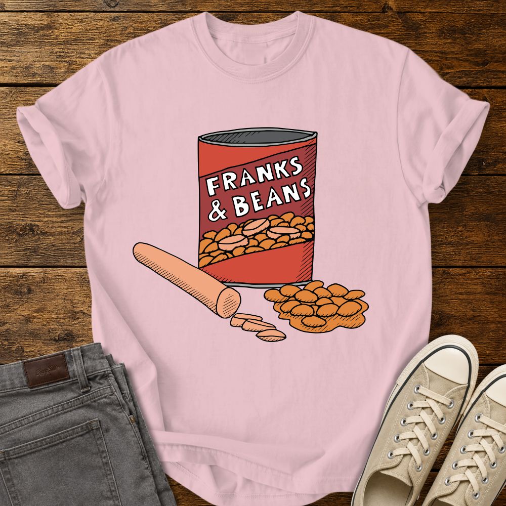 blossom pink graphic T-shirt mockup featuring a humorous illustration of a can labeled “Franks & Beans” with sliced hot dogs and beans, styled with gray jeans and beige sneakers on a rustic wood background.