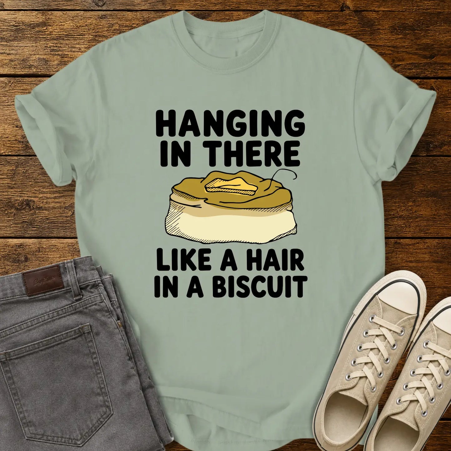 bay green T-shirt mockup featuring the phrase “HANGING IN THERE LIKE A HAIR IN A BISCUIT” in bold black lettering with a hand-drawn biscuit illustration and a single hair resting on top. The shirt is displayed on a rustic wood background alongside folded gray jeans and cream-colored canvas sneakers.
