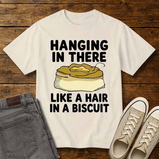Ivory graphic T-shirt mockup featuring the phrase “Hanging In There Like a Hair in a Biscuit” in bold black lettering with a biscuit illustration and a single hair on top, styled with gray jeans and beige sneakers on a rustic wood background.