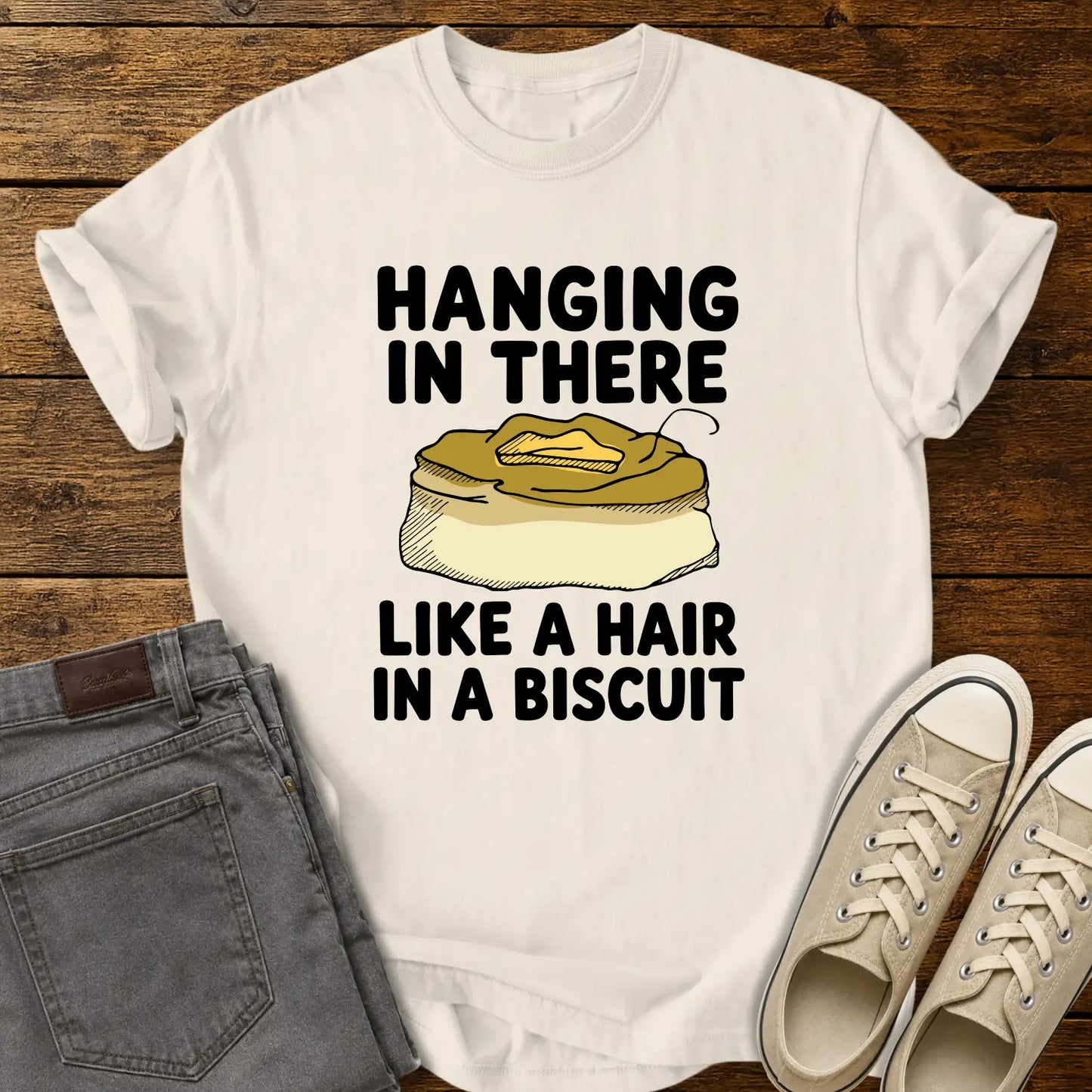 ivory T-shirt mockup featuring the phrase “HANGING IN THERE LIKE A HAIR IN A BISCUIT” in bold black lettering with a hand-drawn biscuit illustration and a single hair resting on top. The shirt is displayed on a rustic wood background alongside folded gray jeans and cream-colored canvas sneakers.