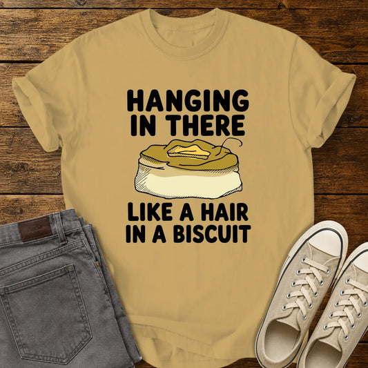 Mustard yellow T-shirt mockup featuring the phrase “HANGING IN THERE LIKE A HAIR IN A BISCUIT” in bold black lettering with a hand-drawn biscuit illustration and a single hair resting on top. The shirt is displayed on a rustic wood background alongside folded gray jeans and cream-colored canvas sneakers.