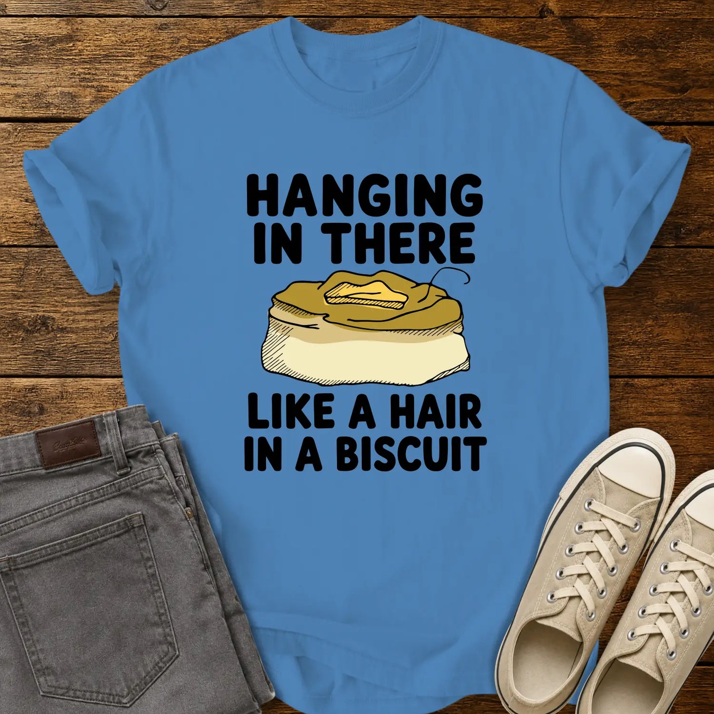 royal caribe blue T-shirt mockup featuring the phrase “HANGING IN THERE LIKE A HAIR IN A BISCUIT” in bold black lettering with a hand-drawn biscuit illustration and a single hair resting on top. The shirt is displayed on a rustic wood background alongside folded gray jeans and cream-colored canvas sneakers.