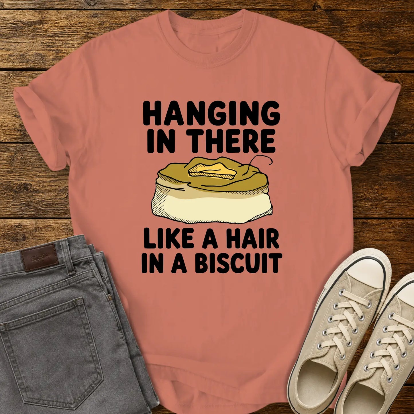 terracotta T-shirt mockup featuring the phrase “HANGING IN THERE LIKE A HAIR IN A BISCUIT” in bold black lettering with a hand-drawn biscuit illustration and a single hair resting on top. The shirt is displayed on a rustic wood background alongside folded gray jeans and cream-colored canvas sneakers.