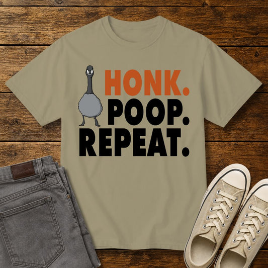 Khaki graphic T-shirt mockup featuring a goose illustration and the phrase “Honk. Poop. Repeat.” in bold orange and black lettering, styled with gray jeans and beige sneakers on a rustic wood background.