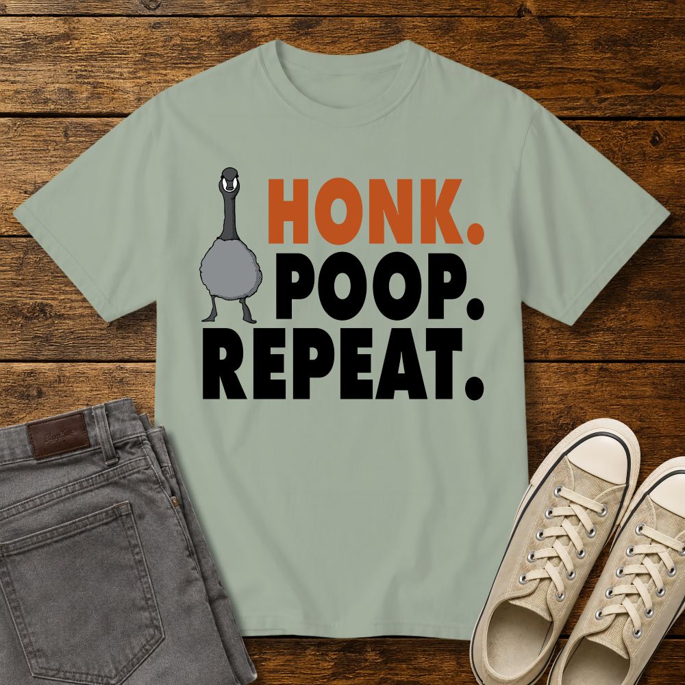 sage green graphic T-shirt mockup featuring a goose illustration and the phrase “Honk. Poop. Repeat.” in bold orange and black lettering, styled with gray jeans and beige sneakers on a rustic wood background.