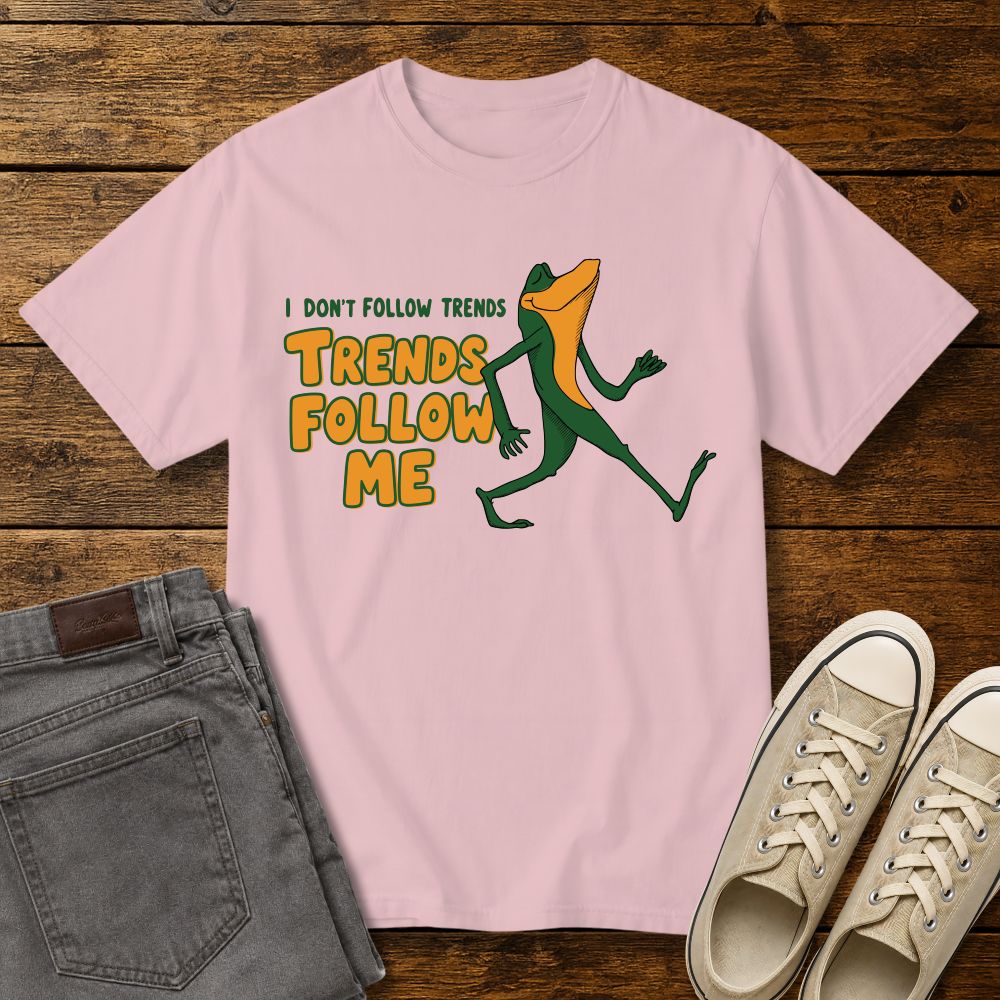 blossom pink graphic T-shirt mockup featuring a cartoon lizard walking confidently beside the phrase “I Don’t Follow Trends, Trends Follow Me” in retro-style green and orange lettering, styled with gray jeans and beige sneakers on a rustic wood background.