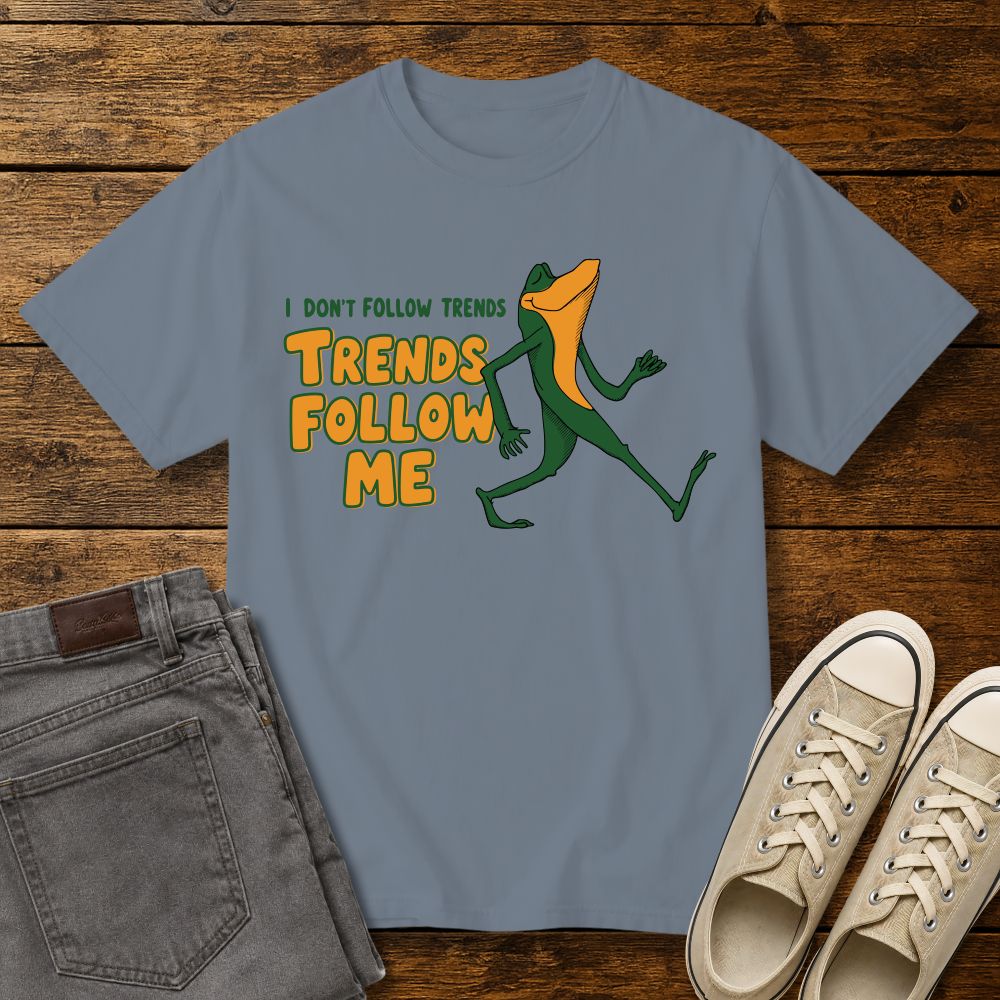 blue jean graphic T-shirt mockup featuring a cartoon lizard walking confidently beside the phrase “I Don’t Follow Trends, Trends Follow Me” in retro-style green and orange lettering, styled with gray jeans and beige sneakers on a rustic wood background.