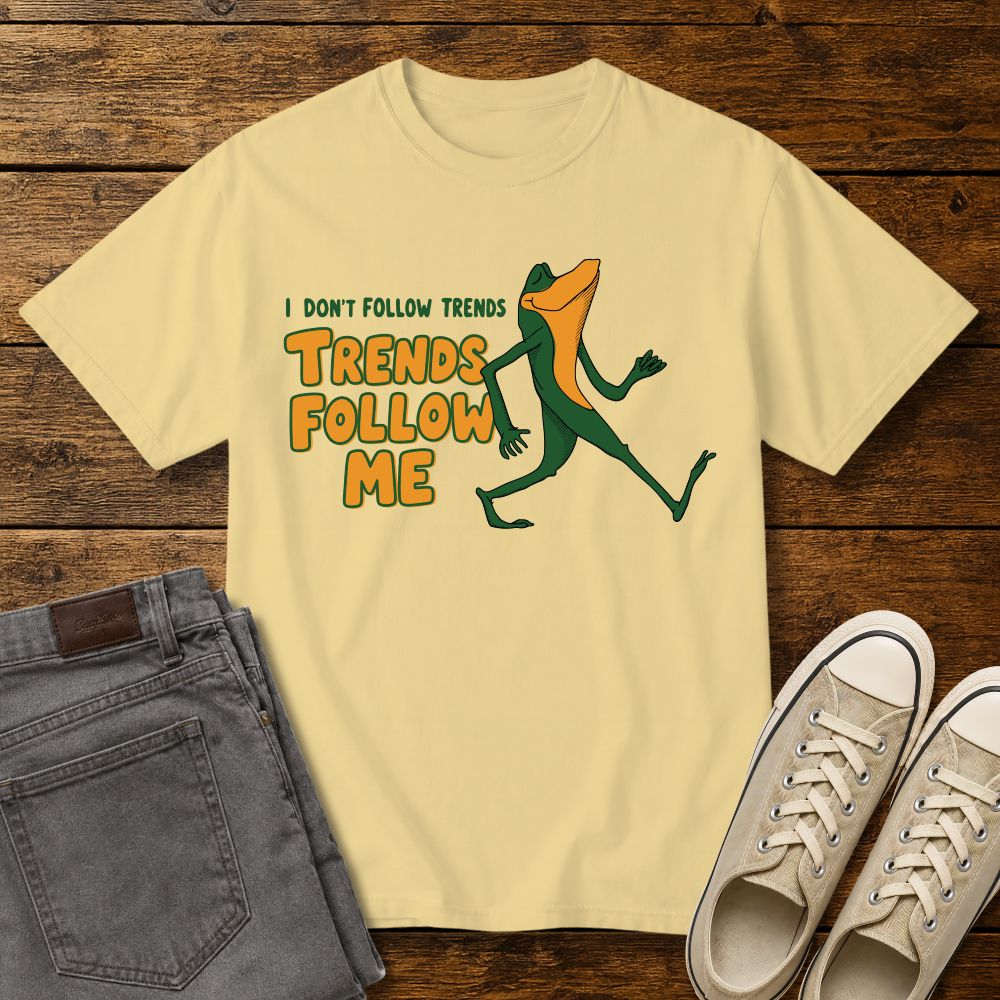 butter color graphic T-shirt mockup featuring a cartoon lizard walking confidently beside the phrase “I Don’t Follow Trends, Trends Follow Me” in retro-style green and orange lettering, styled with gray jeans and beige sneakers on a rustic wood background.