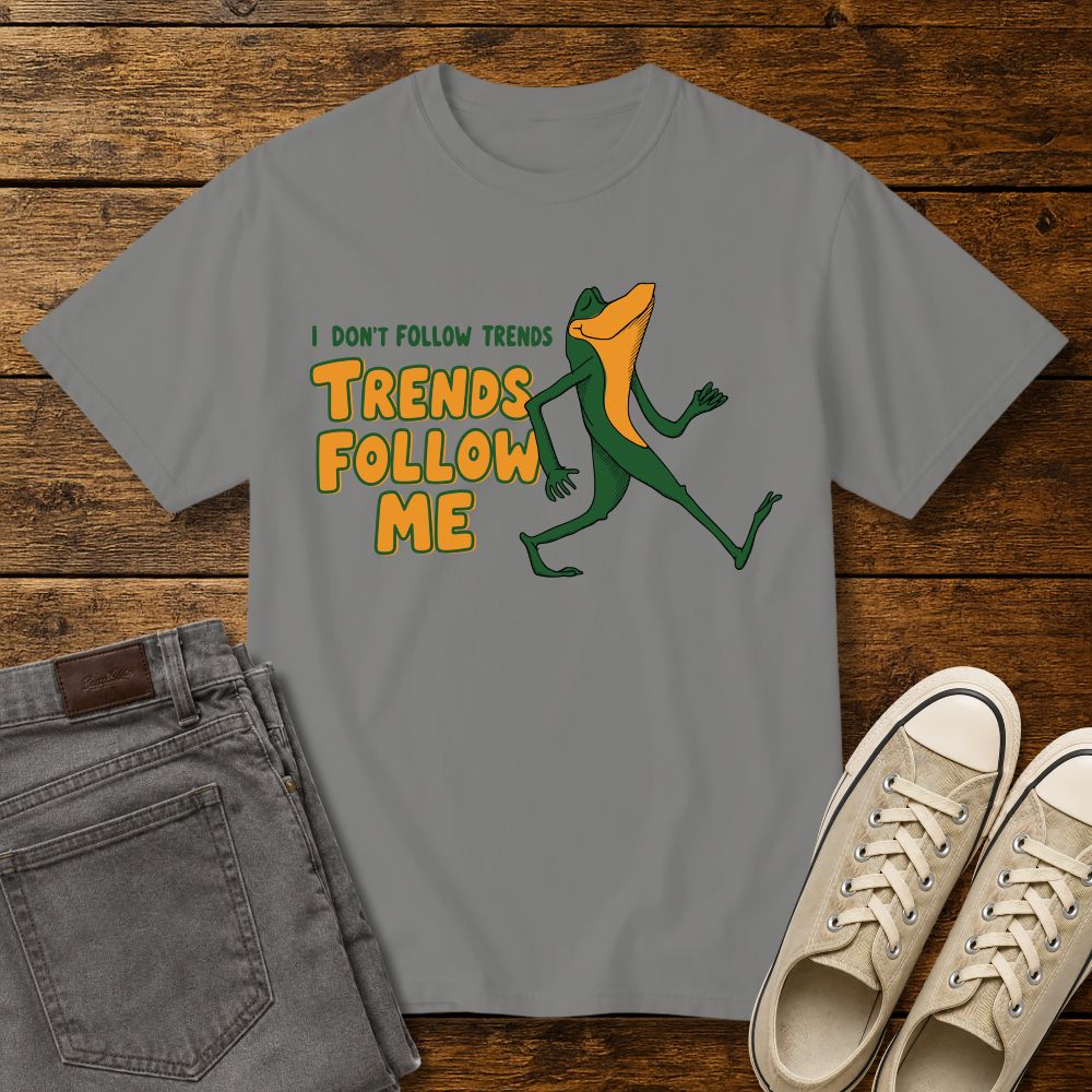 granite graphic T-shirt mockup featuring a cartoon lizard walking confidently beside the phrase “I Don’t Follow Trends, Trends Follow Me” in retro-style green and orange lettering, styled with gray jeans and beige sneakers on a rustic wood background.