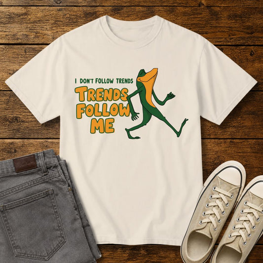 Ivory graphic T-shirt mockup featuring a cartoon lizard walking confidently beside the phrase “I Don’t Follow Trends, Trends Follow Me” in retro-style green and orange lettering, styled with gray jeans and beige sneakers on a rustic wood background.