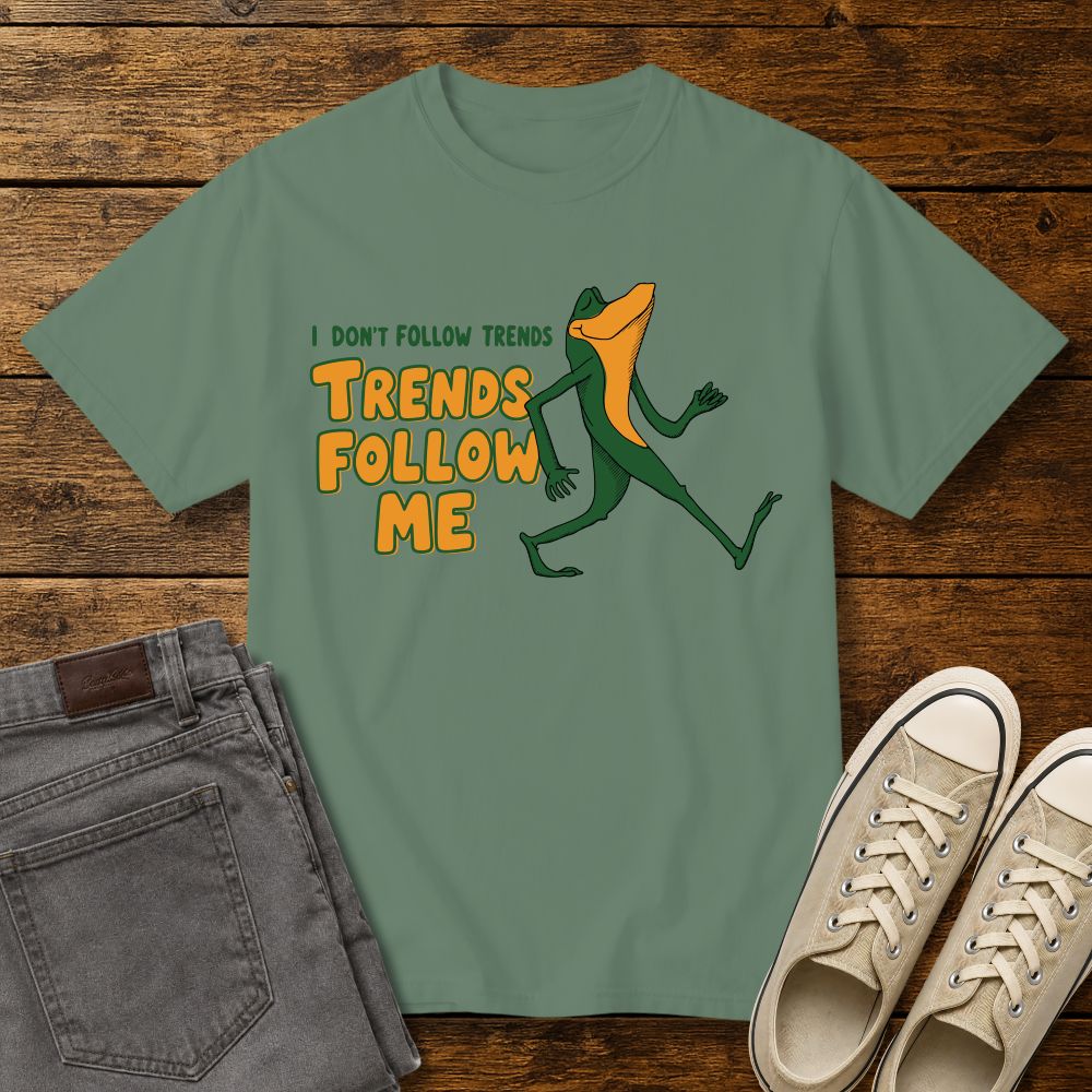 light green graphic T-shirt mockup featuring a cartoon lizard walking confidently beside the phrase “I Don’t Follow Trends, Trends Follow Me” in retro-style green and orange lettering, styled with gray jeans and beige sneakers on a rustic wood background.