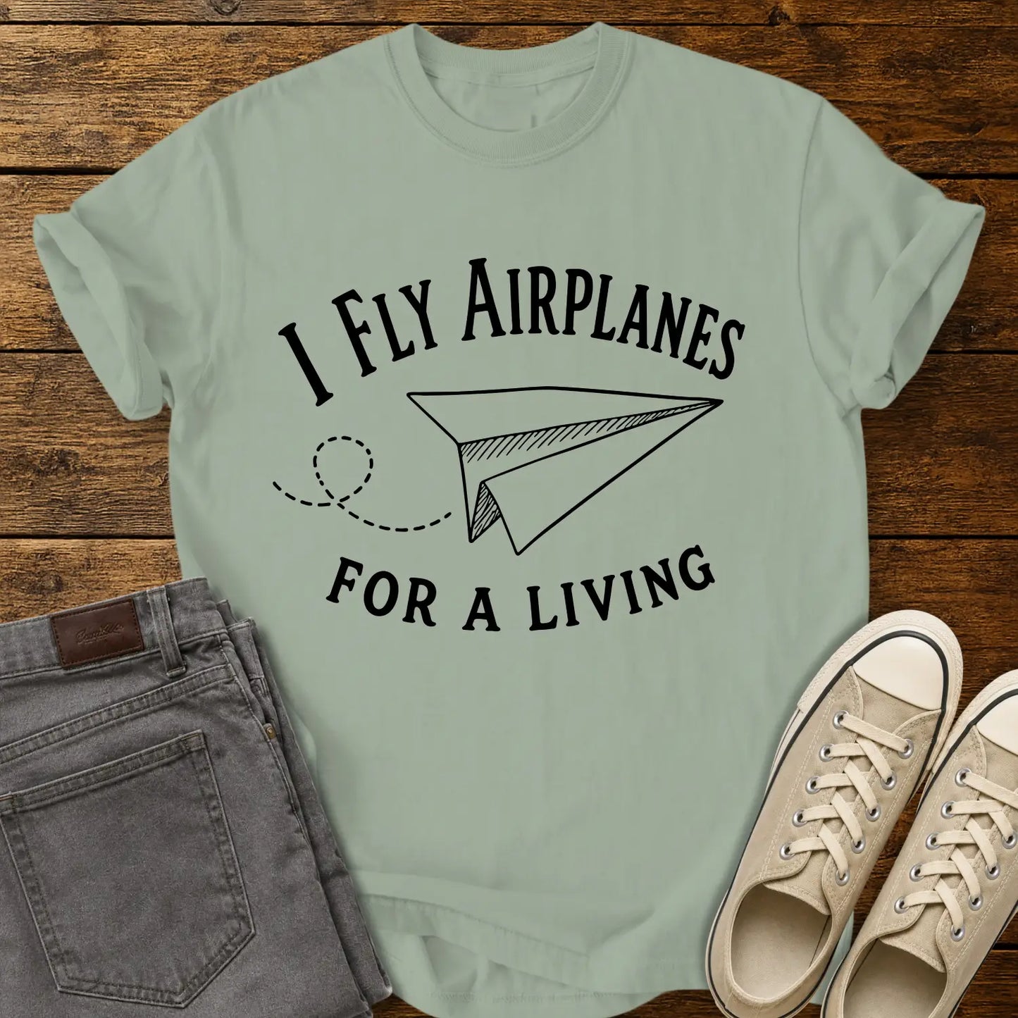 bay green T-shirt mockup featuring the phrase “I FLY AIRPLANES FOR A LIVING” in bold vintage lettering surrounding a hand-drawn paper airplane with a looping flight path. The shirt is displayed on a rustic wood plank background alongside gray jeans and cream canvas sneakers, creating a casual retro-inspired apparel presentation.