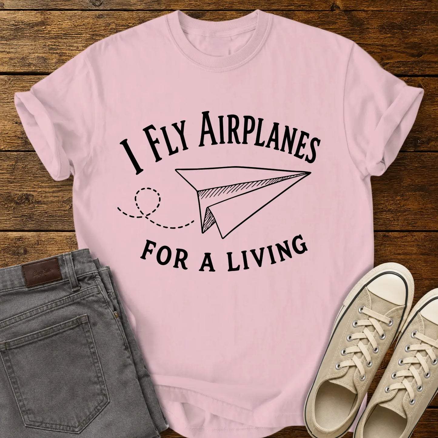 blossom pink T-shirt mockup featuring the phrase “I FLY AIRPLANES FOR A LIVING” in bold vintage lettering surrounding a hand-drawn paper airplane with a looping flight path. The shirt is displayed on a rustic wood plank background alongside gray jeans and cream canvas sneakers, creating a casual retro-inspired apparel presentation.