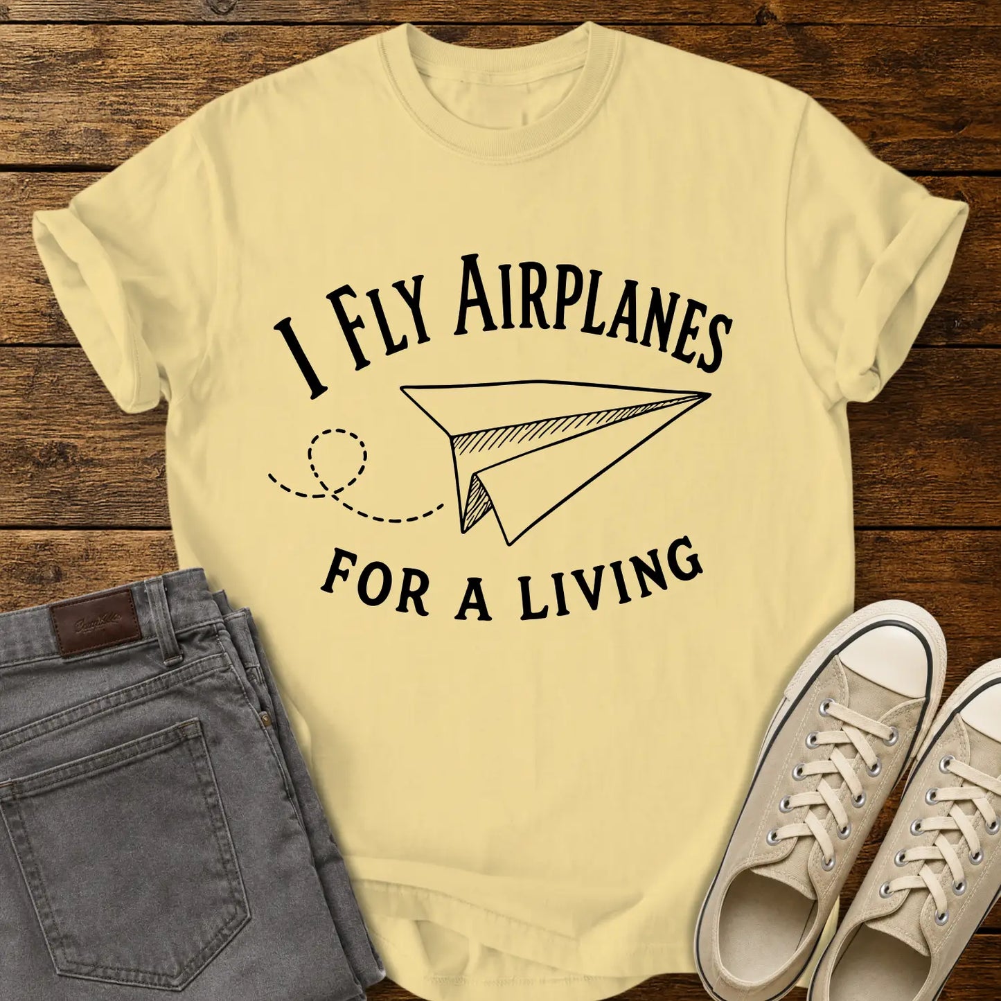 butter yellow T-shirt mockup featuring the phrase “I FLY AIRPLANES FOR A LIVING” in bold vintage lettering surrounding a hand-drawn paper airplane with a looping flight path. The shirt is displayed on a rustic wood plank background alongside gray jeans and cream canvas sneakers, creating a casual retro-inspired apparel presentation.