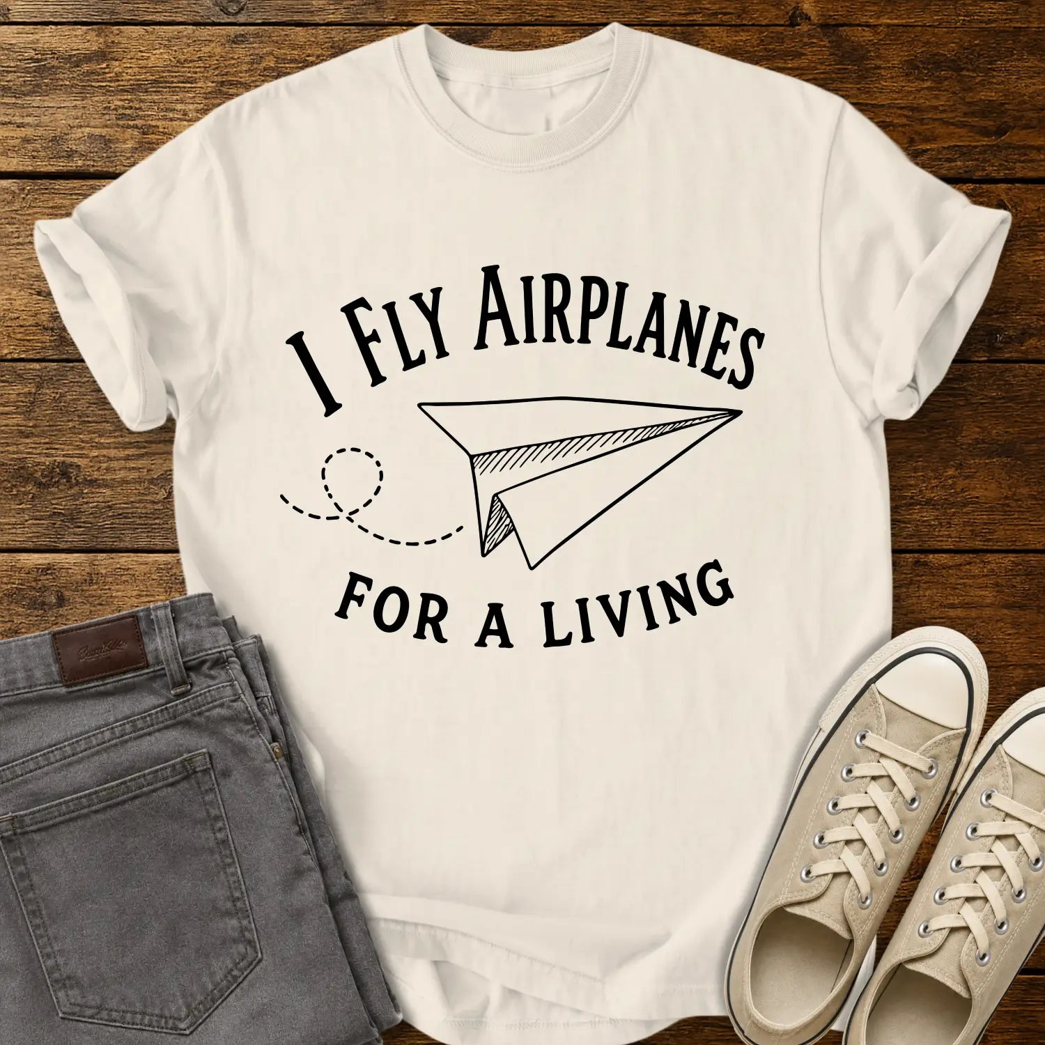 Ivory T-shirt mockup featuring the phrase “I FLY AIRPLANES FOR A LIVING” in bold vintage lettering surrounding a hand-drawn paper airplane with a looping flight path. The shirt is displayed on a rustic wood plank background alongside gray jeans and cream canvas sneakers, creating a casual retro-inspired apparel presentation.