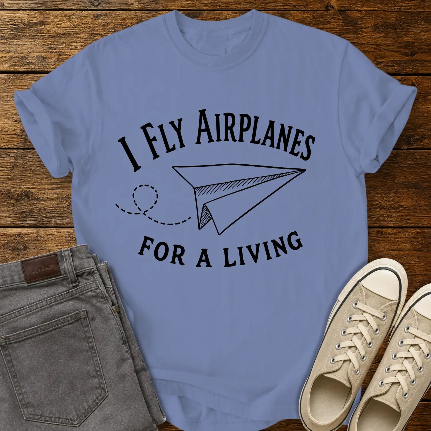 washed denim blue T-shirt mockup featuring the phrase “I FLY AIRPLANES FOR A LIVING” in bold vintage lettering surrounding a hand-drawn paper airplane with a looping flight path. The shirt is displayed on a rustic wood plank background alongside gray jeans and cream canvas sneakers, creating a casual retro-inspired apparel presentation.
