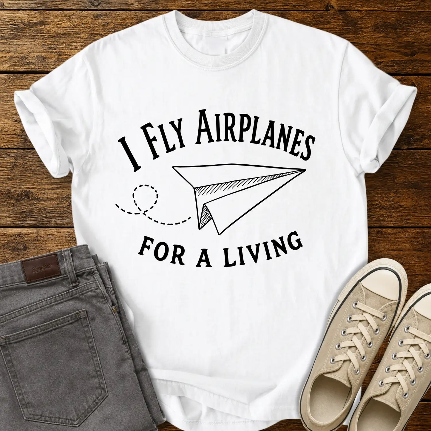 white T-shirt mockup featuring the phrase “I FLY AIRPLANES FOR A LIVING” in bold vintage lettering surrounding a hand-drawn paper airplane with a looping flight path. The shirt is displayed on a rustic wood plank background alongside gray jeans and cream canvas sneakers, creating a casual retro-inspired apparel presentation.