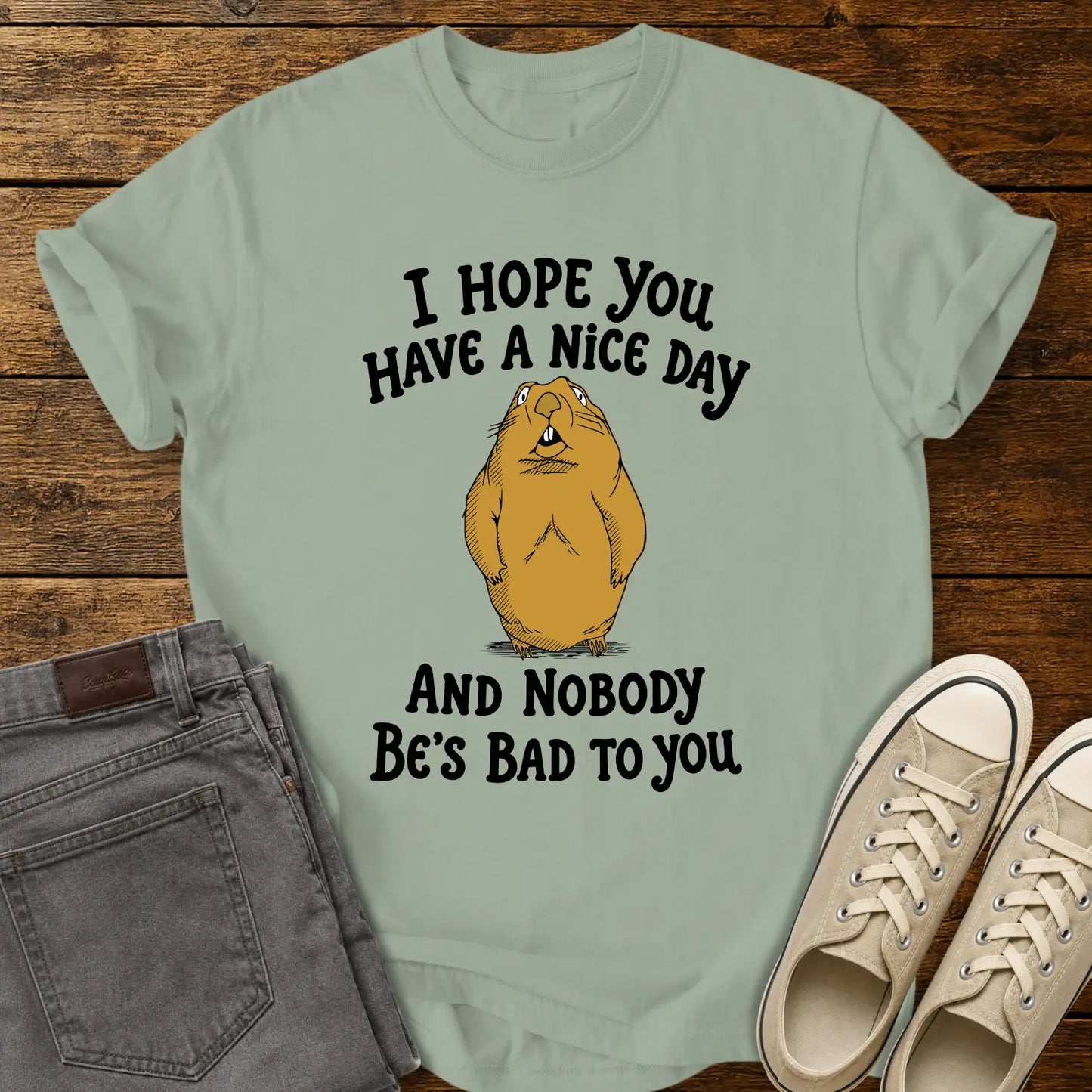bay green T-shirt featuring a cartoon groundhog beneath the phrase “I HOPE YOU HAVE A NICE DAY AND NOBODY BE'S BAD TO YOU.” The shirt is displayed in a flat-lay on a rustic wood plank background with folded gray jeans and cream canvas sneakers.