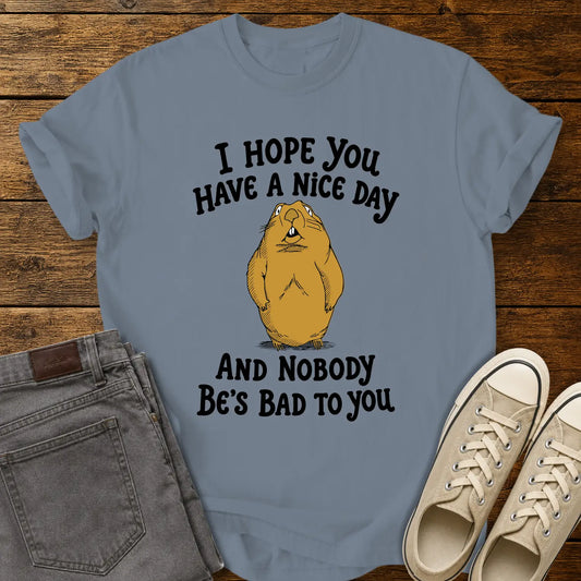 Blue Jean T-shirt featuring a cartoon groundhog beneath the phrase “I HOPE YOU HAVE A NICE DAY AND NOBODY BE'S BAD TO YOU.” The shirt is displayed in a flat-lay on a rustic wood plank background with folded gray jeans and cream canvas sneakers.