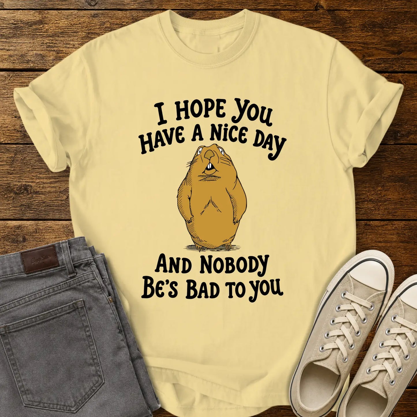 butter T-shirt featuring a cartoon groundhog beneath the phrase “I HOPE YOU HAVE A NICE DAY AND NOBODY BE'S BAD TO YOU.” The shirt is displayed in a flat-lay on a rustic wood plank background with folded gray jeans and cream canvas sneakers.