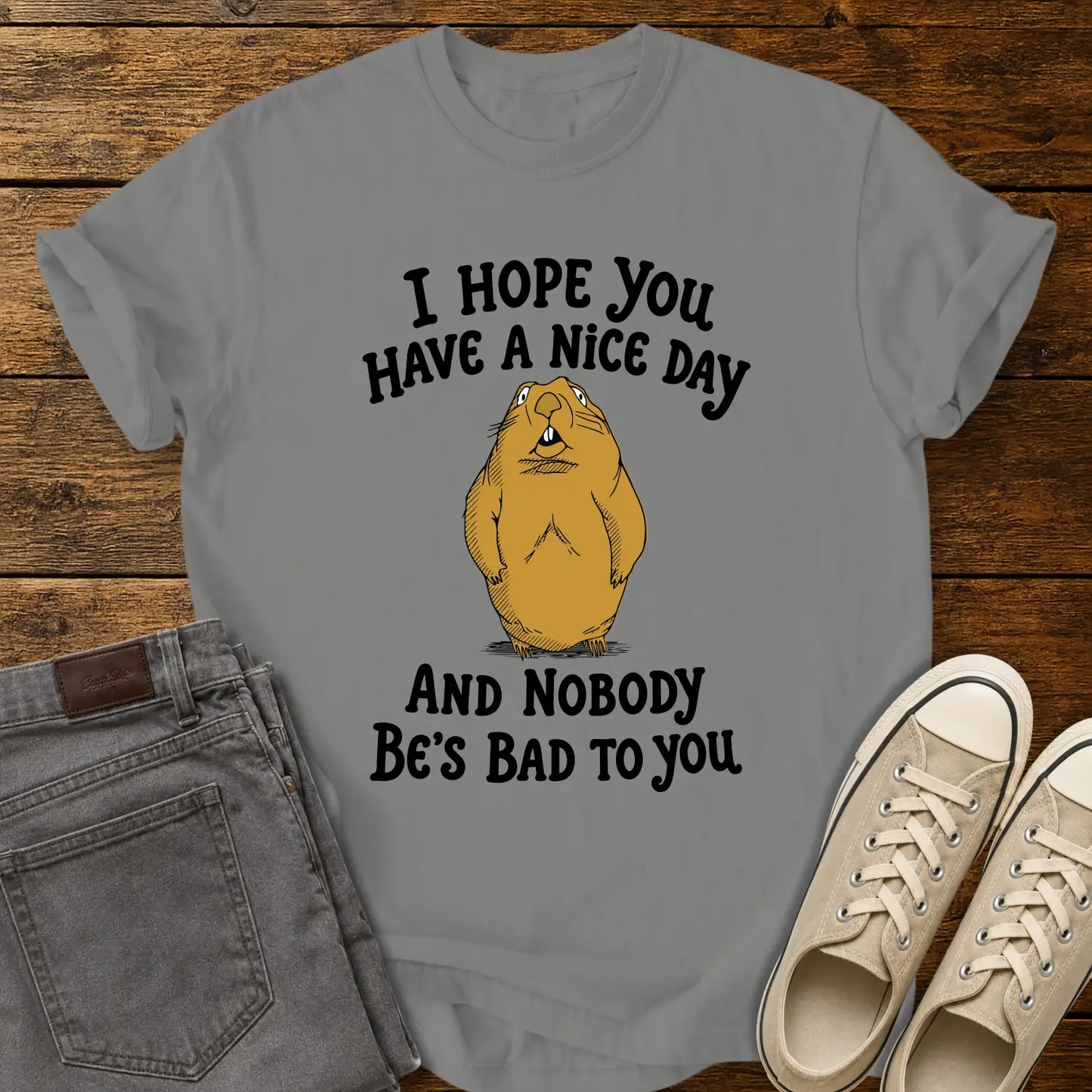 granite T-shirt featuring a cartoon groundhog beneath the phrase “I HOPE YOU HAVE A NICE DAY AND NOBODY BE'S BAD TO YOU.” The shirt is displayed in a flat-lay on a rustic wood plank background with folded gray jeans and cream canvas sneakers.