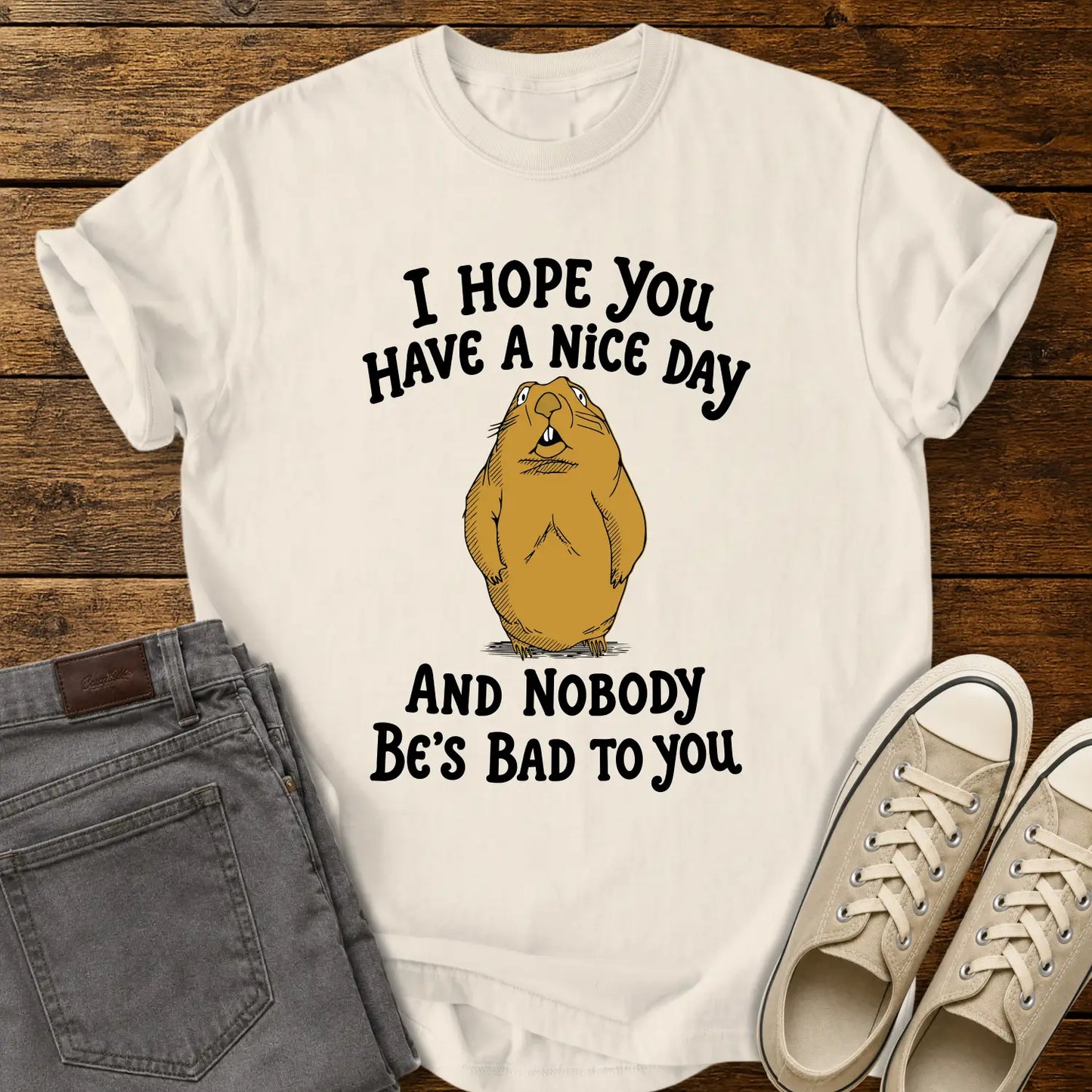 ivory T-shirt featuring a cartoon groundhog beneath the phrase “I HOPE YOU HAVE A NICE DAY AND NOBODY BE'S BAD TO YOU.” The shirt is displayed in a flat-lay on a rustic wood plank background with folded gray jeans and cream canvas sneakers.