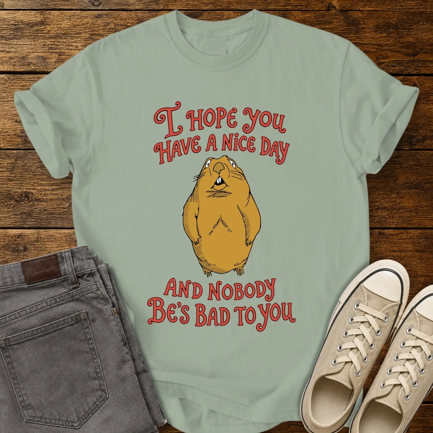 bay green graphic T-shirt mockup featuring a groundhog illustration between the phrases “I Hope You Have a Nice Day” and “And Nobody Be’s Bad to You” in vintage-style red lettering, styled with gray jeans and beige sneakers on a rustic wood background.
