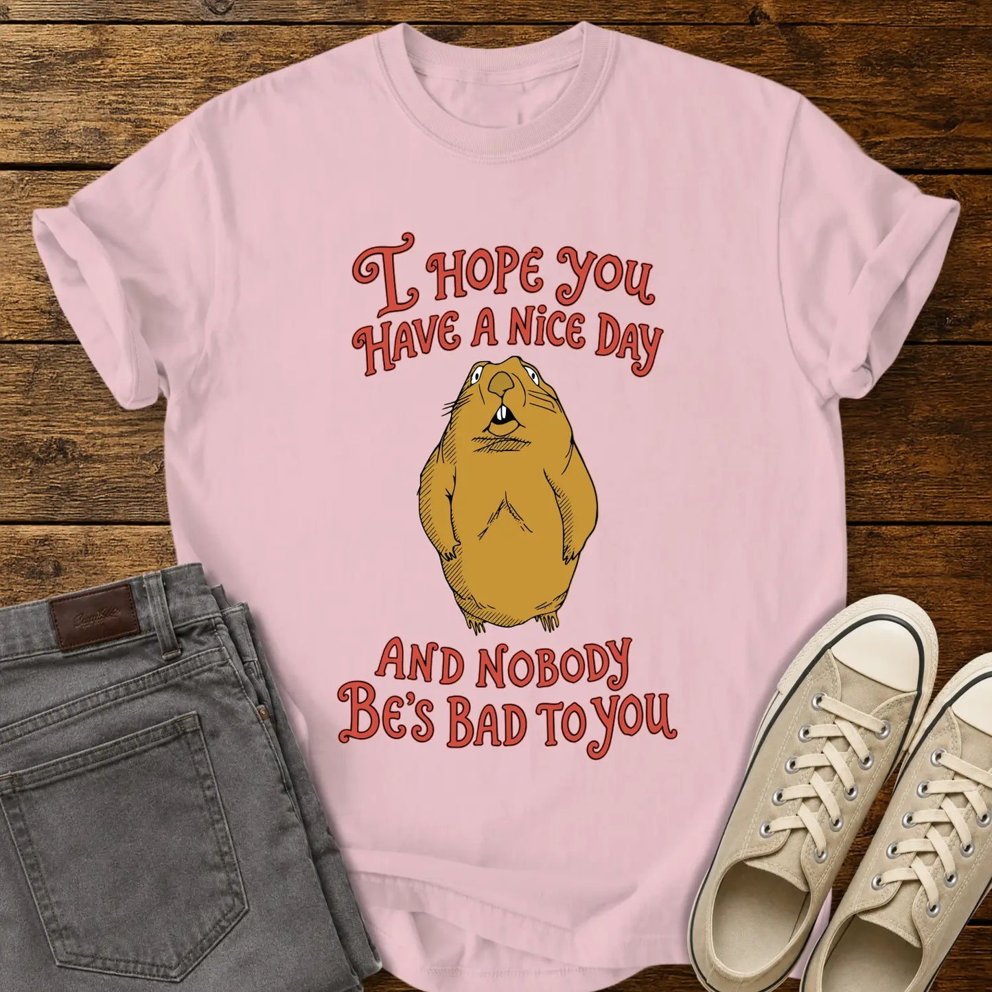 blossom pink graphic T-shirt mockup featuring a groundhog illustration between the phrases “I Hope You Have a Nice Day” and “And Nobody Be’s Bad to You” in vintage-style red lettering, styled with gray jeans and beige sneakers on a rustic wood background.