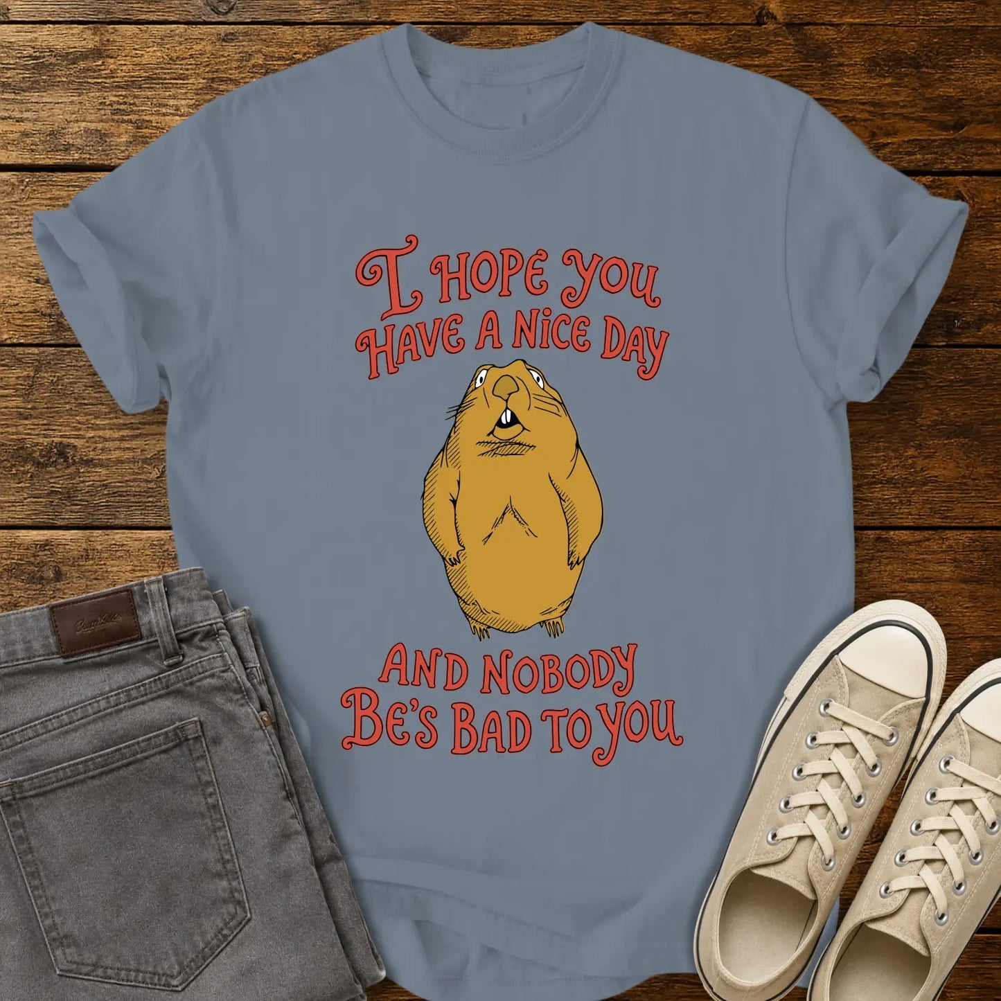 blue jean graphic T-shirt mockup featuring a groundhog illustration between the phrases “I Hope You Have a Nice Day” and “And Nobody Be’s Bad to You” in vintage-style red lettering, styled with gray jeans and beige sneakers on a rustic wood background.