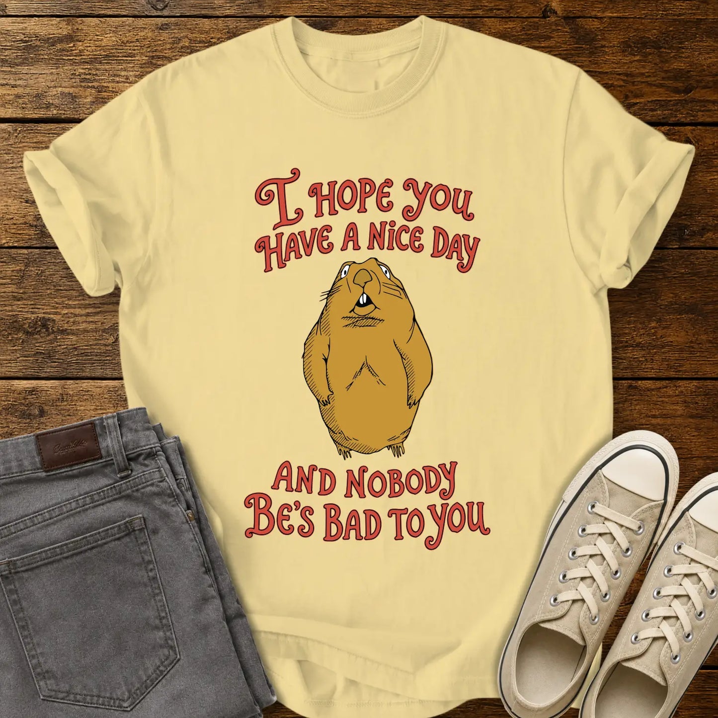 butter yellow graphic T-shirt mockup featuring a groundhog illustration between the phrases “I Hope You Have a Nice Day” and “And Nobody Be’s Bad to You” in vintage-style red lettering, styled with gray jeans and beige sneakers on a rustic wood background.