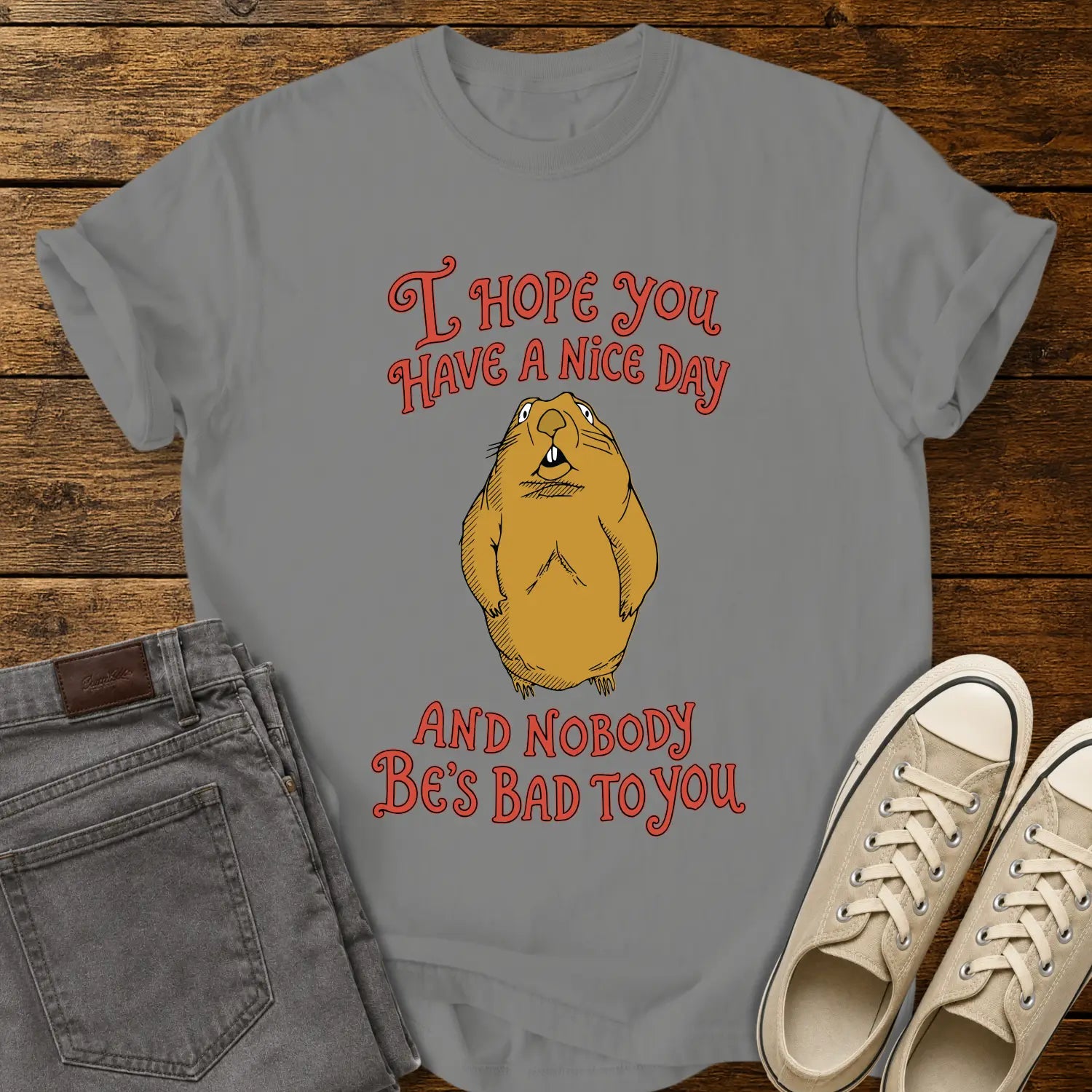 Granite gray graphic T-shirt mockup featuring a groundhog illustration between the phrases “I Hope You Have a Nice Day” and “And Nobody Be’s Bad to You” in vintage-style red lettering, styled with gray jeans and beige sneakers on a rustic wood background.