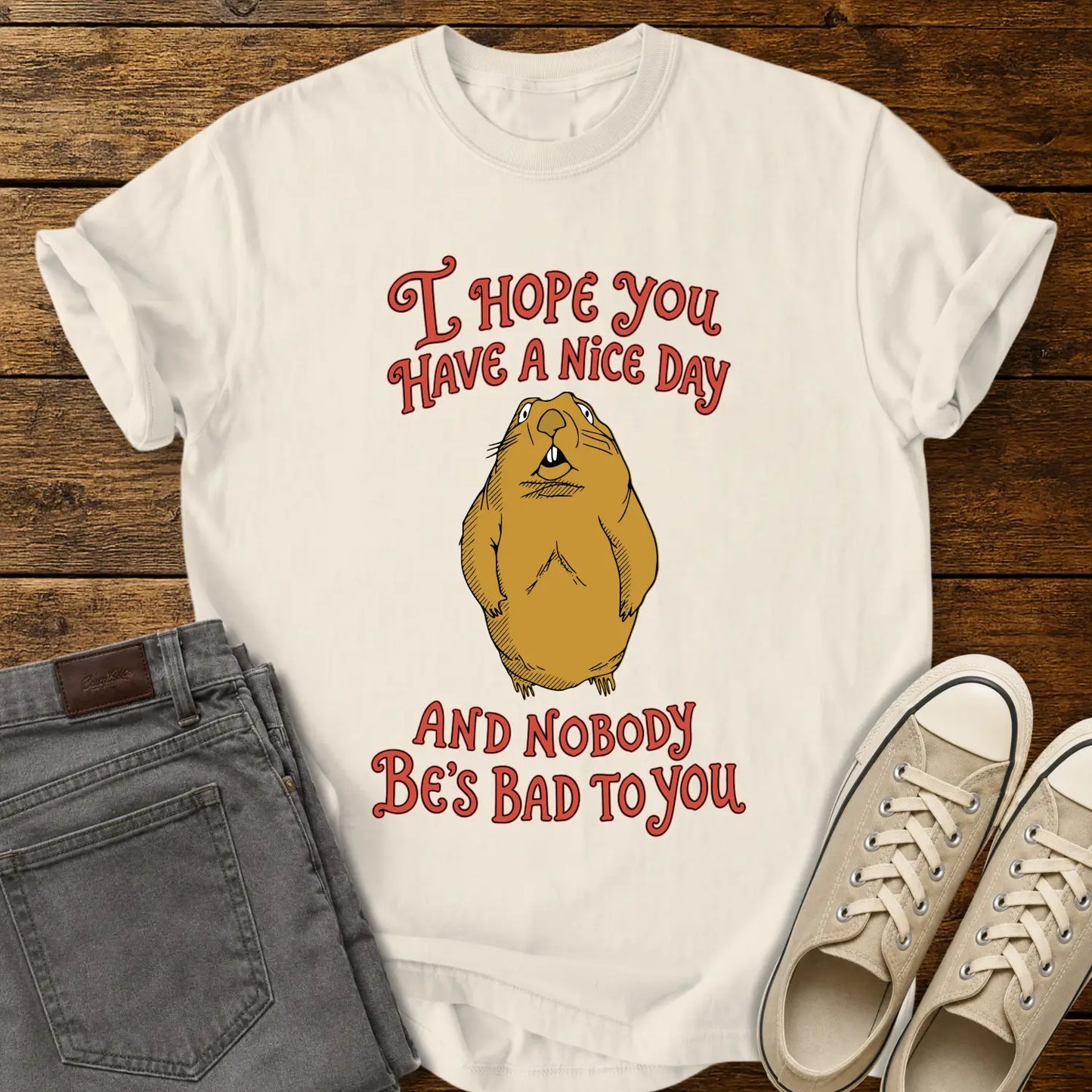 ivory graphic T-shirt mockup featuring a groundhog illustration between the phrases “I Hope You Have a Nice Day” and “And Nobody Be’s Bad to You” in vintage-style red lettering, styled with gray jeans and beige sneakers on a rustic wood background.
