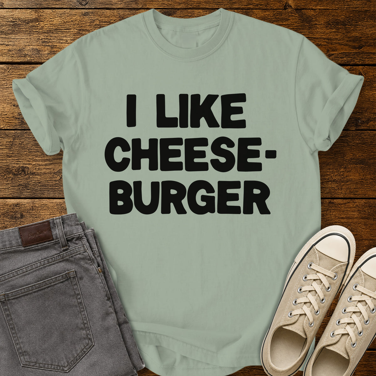 bay green graphic T-shirt featuring bold black lettering reading “I Like Cheeseburger” in a playful retro font, displayed on a rustic wooden background with folded gray jeans and beige canvas sneakers.