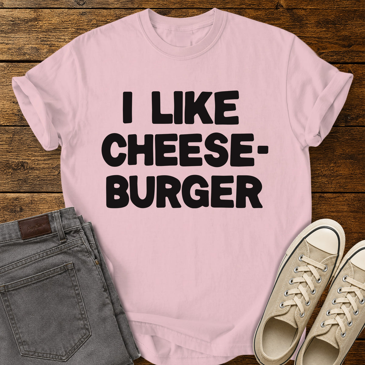blossom pink graphic T-shirt featuring bold black lettering reading “I Like Cheeseburger” in a playful retro font, displayed on a rustic wooden background with folded gray jeans and beige canvas sneakers.