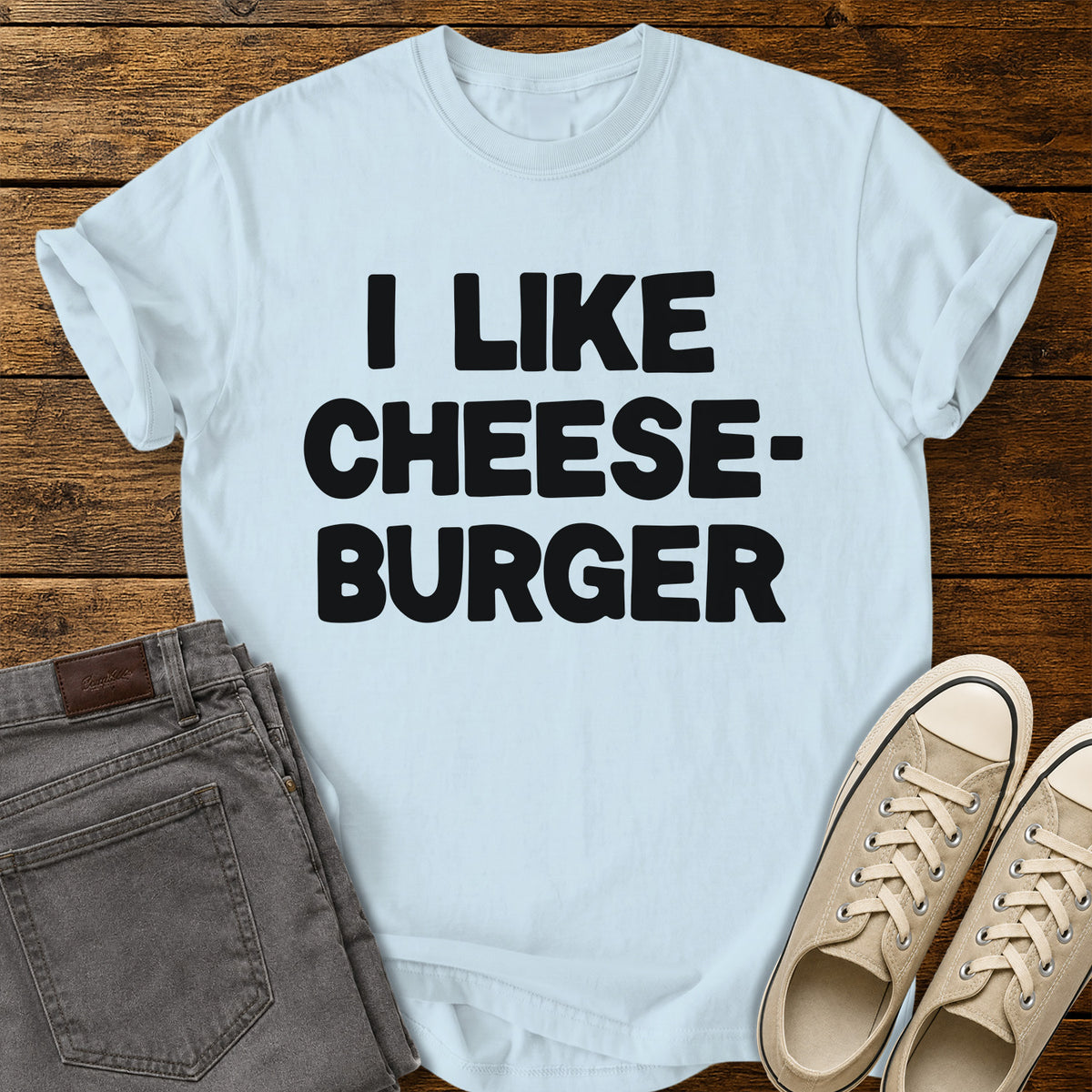 chambray blue graphic T-shirt featuring bold black lettering reading “I Like Cheeseburger” in a playful retro font, displayed on a rustic wooden background with folded gray jeans and beige canvas sneakers.