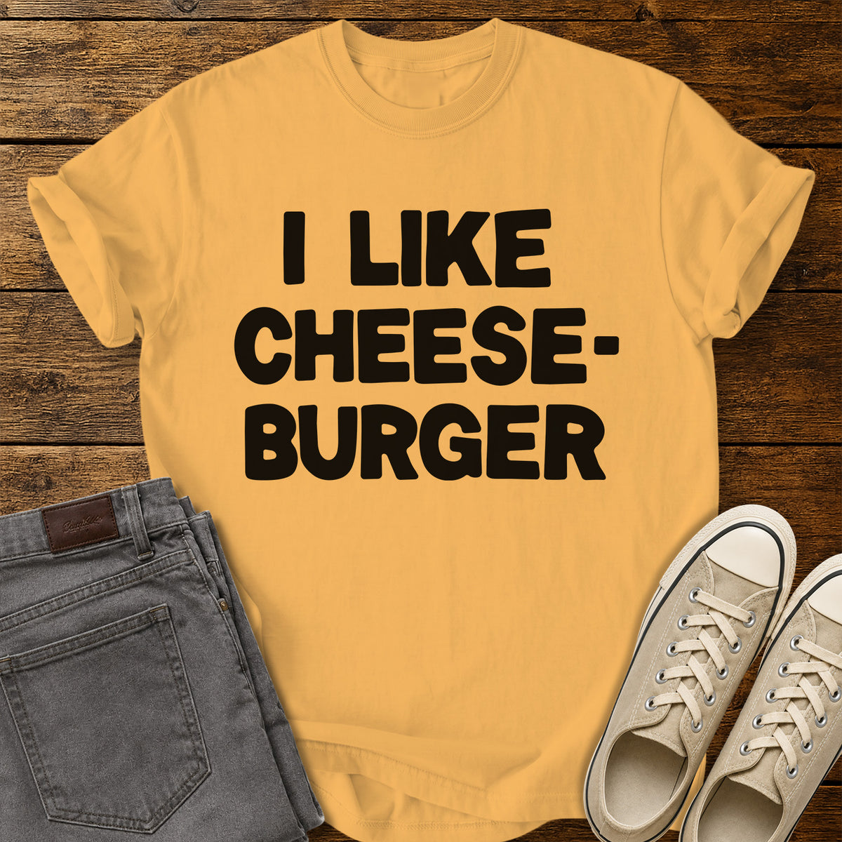 citrus yellow graphic T-shirt featuring bold black lettering reading “I Like Cheeseburger” in a playful retro font, displayed on a rustic wooden background with folded gray jeans and beige canvas sneakers.