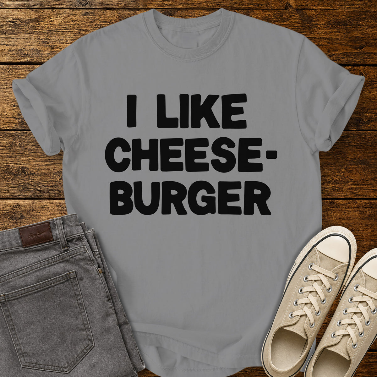 grey graphic T-shirt featuring bold black lettering reading “I Like Cheeseburger” in a playful retro font, displayed on a rustic wooden background with folded gray jeans and beige canvas sneakers.