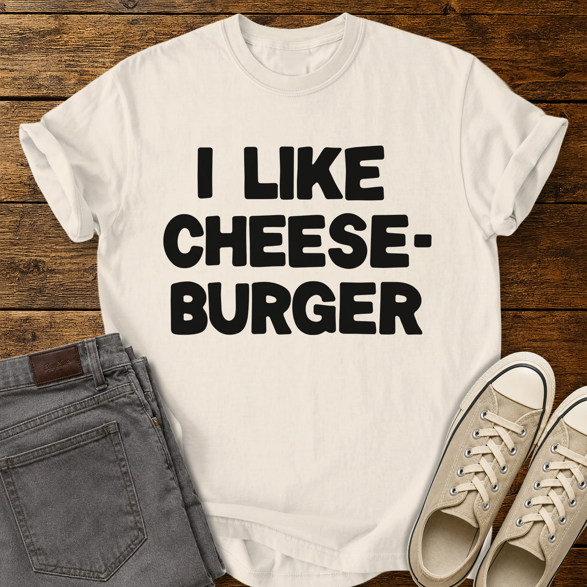 ivory graphic T-shirt featuring bold black lettering reading “I Like Cheeseburger” in a playful retro font, displayed on a rustic wooden background with folded gray jeans and beige canvas sneakers.