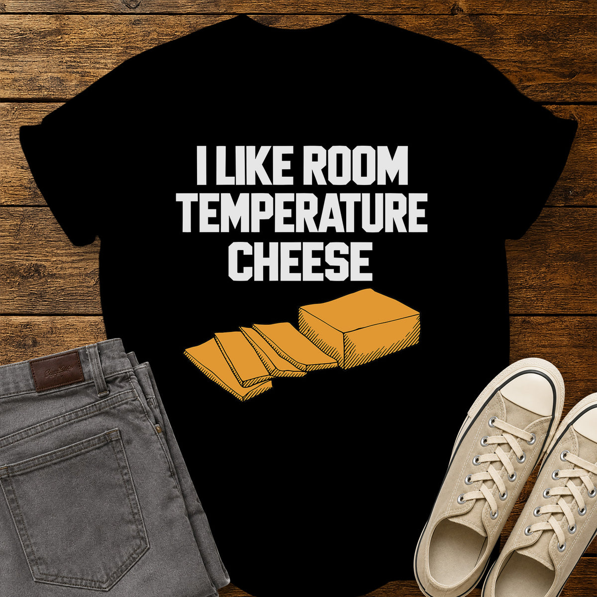 black graphic T-shirt featuring bold white lettering reading “I Like Room Temperature Cheese” above an orange illustration of a block of cheese with several slices, displayed on a rustic wooden background with gray jeans and beige sneakers.