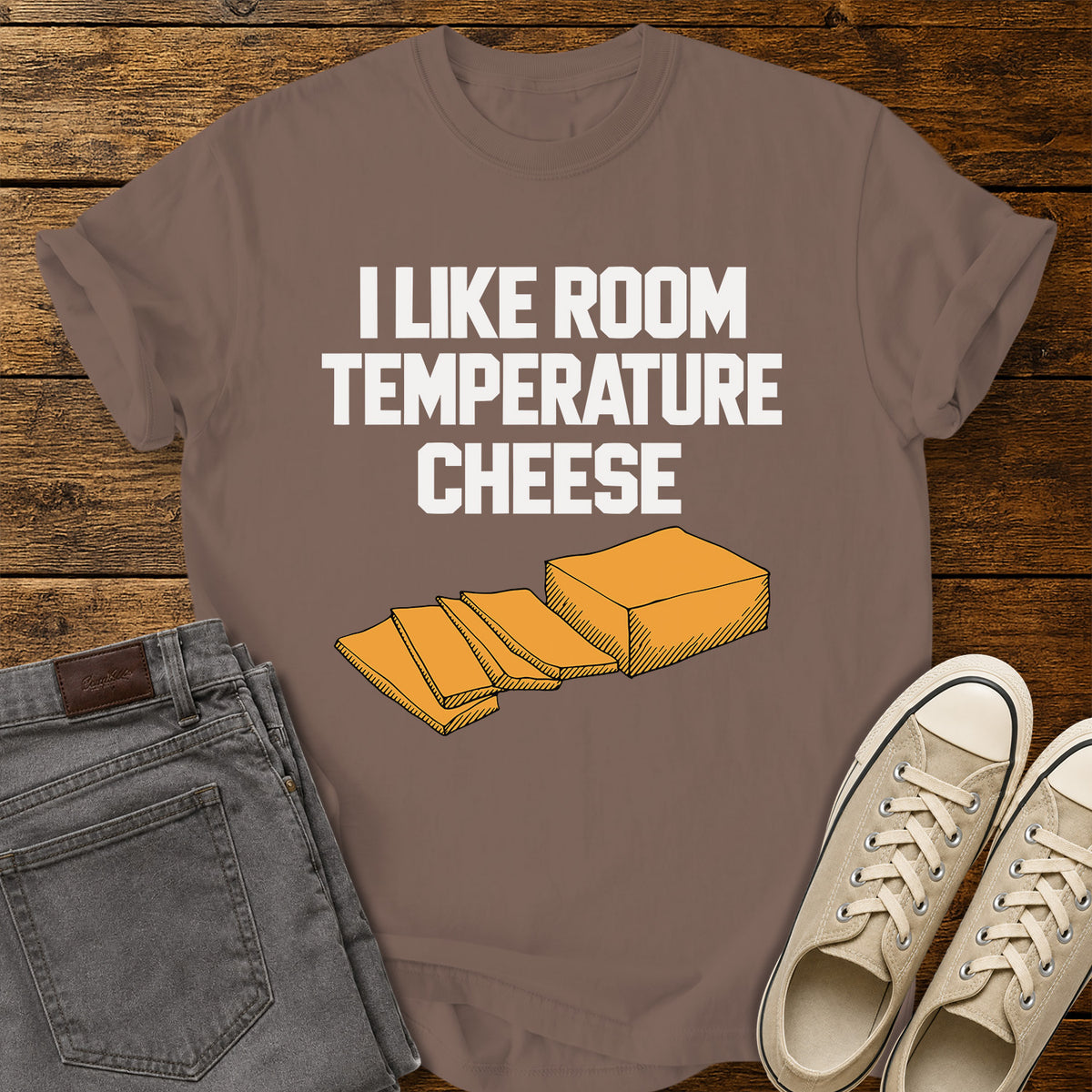 espresso brown graphic T-shirt featuring bold white lettering reading “I Like Room Temperature Cheese” above an orange illustration of a block of cheese with several slices, displayed on a rustic wooden background with gray jeans and beige sneakers.
