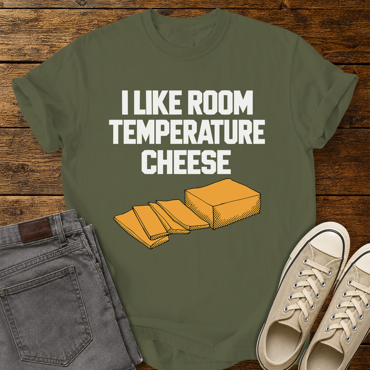 hemp green graphic T-shirt featuring bold white lettering reading “I Like Room Temperature Cheese” above an orange illustration of a block of cheese with several slices, displayed on a rustic wooden background with gray jeans and beige sneakers.