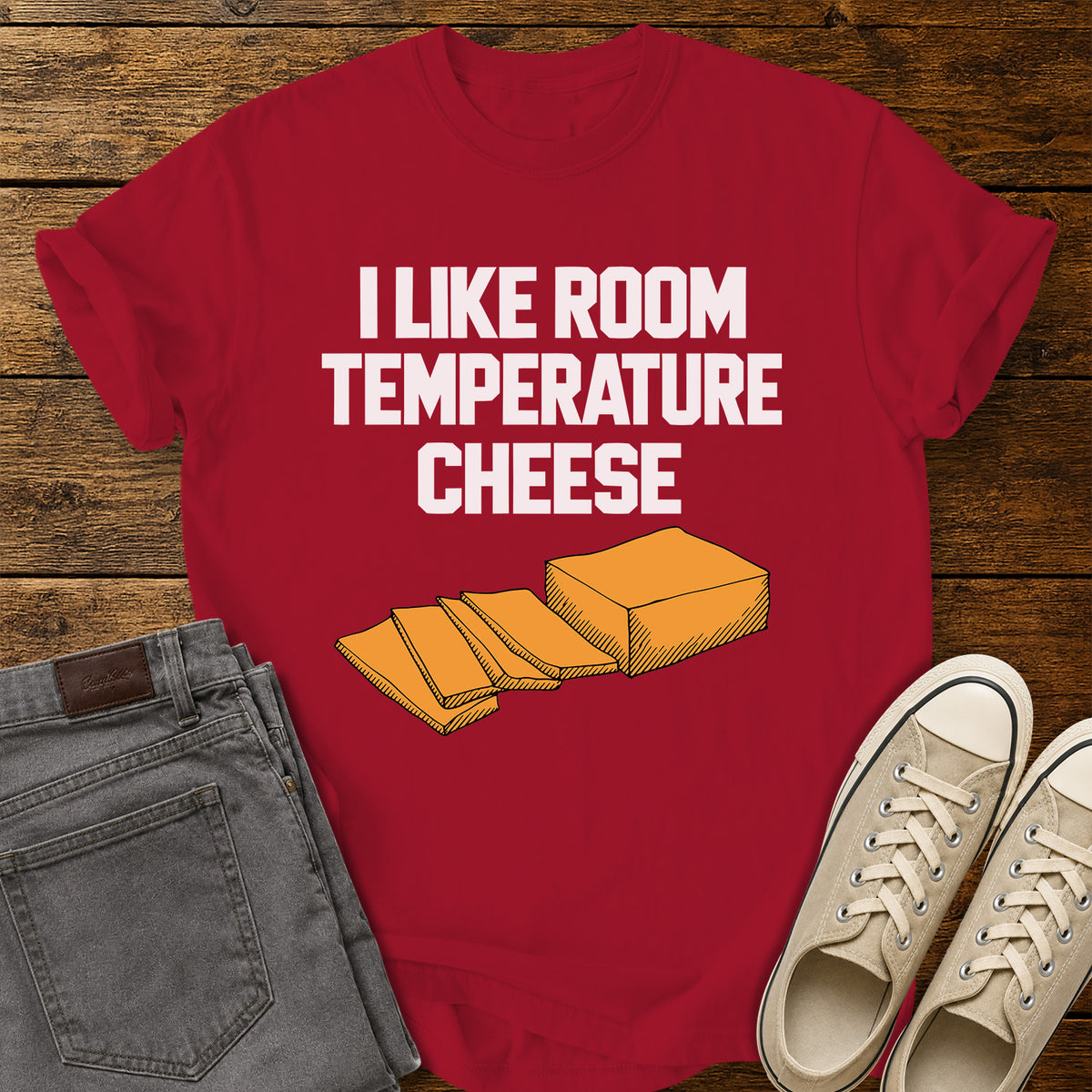 red graphic T-shirt featuring bold white lettering reading “I Like Room Temperature Cheese” above an orange illustration of a block of cheese with several slices, displayed on a rustic wooden background with gray jeans and beige sneakers.