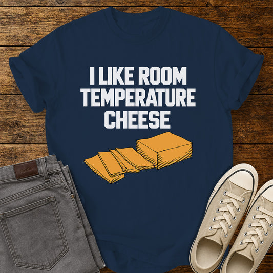 true navy blue graphic T-shirt featuring bold white lettering reading “I Like Room Temperature Cheese” above an orange illustration of a block of cheese with several slices, displayed on a rustic wooden background with gray jeans and beige sneakers.