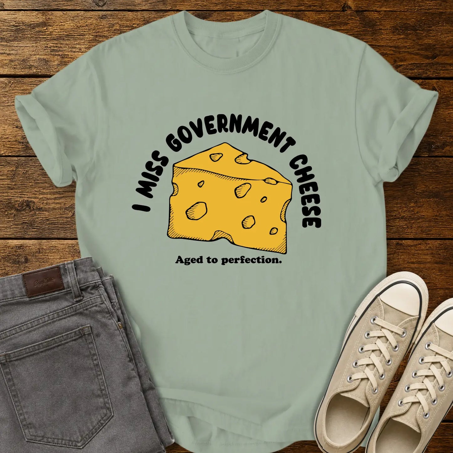 bay green T-shirt mockup featuring a hand-drawn wedge of yellow cheese with the phrase “I MISS GOVERNMENT CHEESE” arched above and “Aged to perfection.” below. The shirt is displayed on a rustic wood background alongside folded gray jeans and cream-colored canvas sneakers.