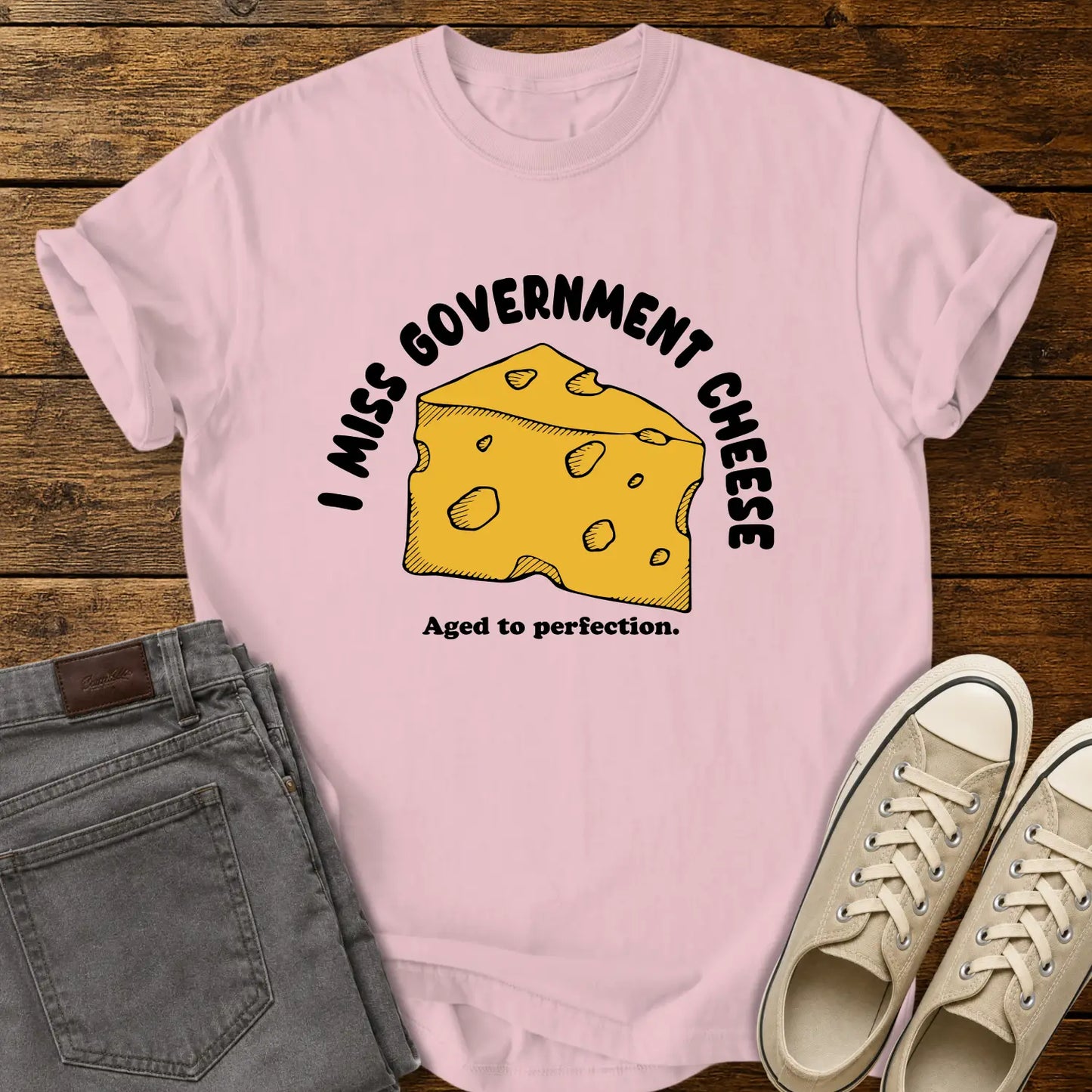 blossom pink T-shirt mockup featuring a hand-drawn wedge of yellow cheese with the phrase “I MISS GOVERNMENT CHEESE” arched above and “Aged to perfection.” below. The shirt is displayed on a rustic wood background alongside folded gray jeans and cream-colored canvas sneakers.