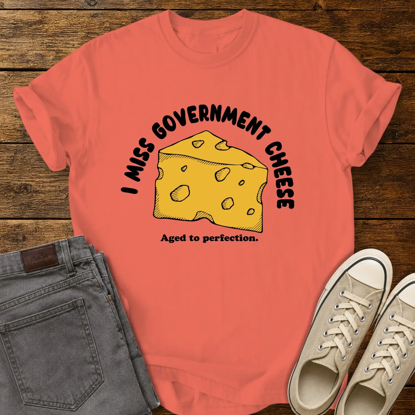 Bright salmon T-shirt mockup featuring a hand-drawn wedge of yellow cheese with the phrase “I MISS GOVERNMENT CHEESE” arched above and “Aged to perfection.” below. The shirt is displayed on a rustic wood background alongside folded gray jeans and cream-colored canvas sneakers.