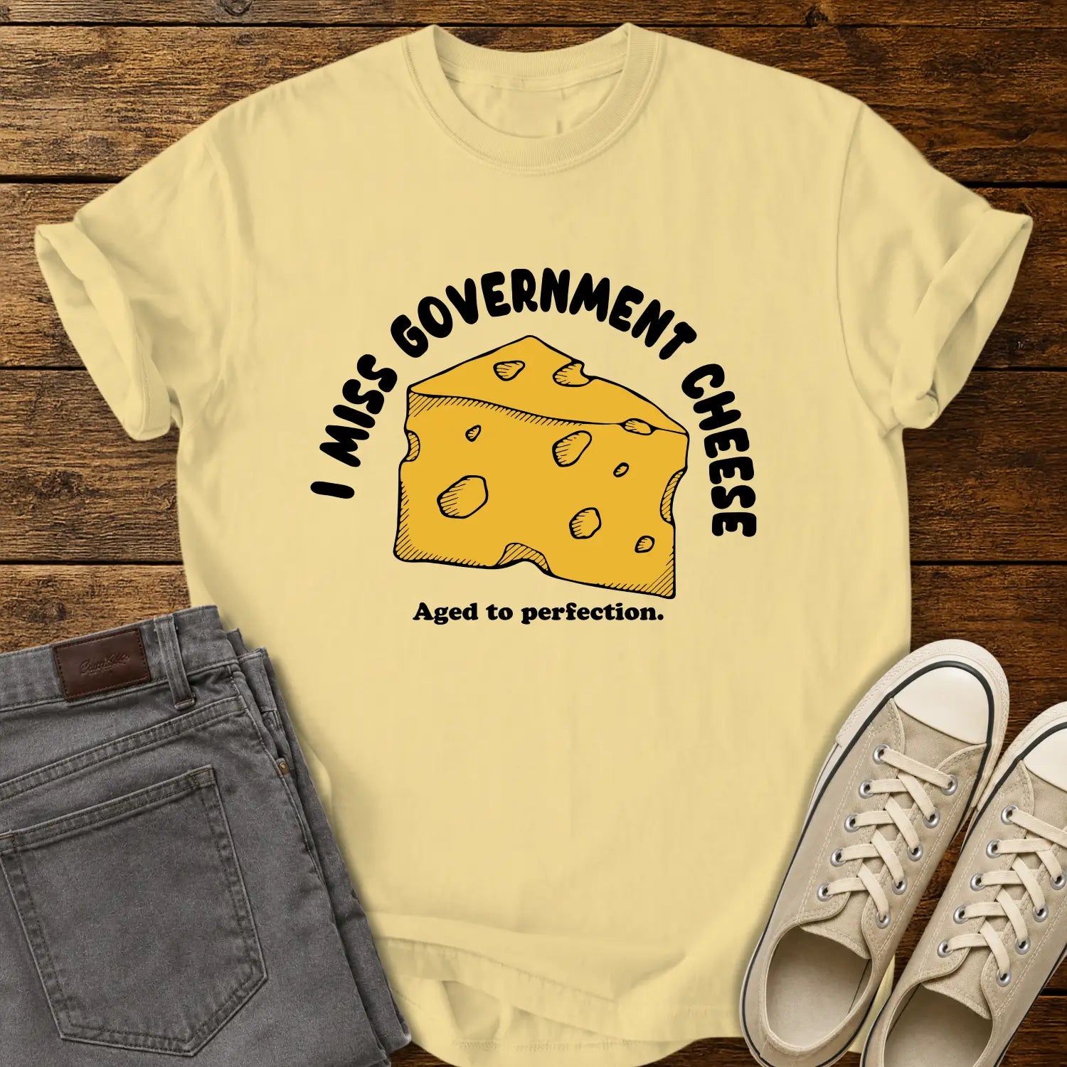 butter yellow T-shirt mockup featuring a hand-drawn wedge of yellow cheese with the phrase “I MISS GOVERNMENT CHEESE” arched above and “Aged to perfection.” below. The shirt is displayed on a rustic wood background alongside folded gray jeans and cream-colored canvas sneakers.