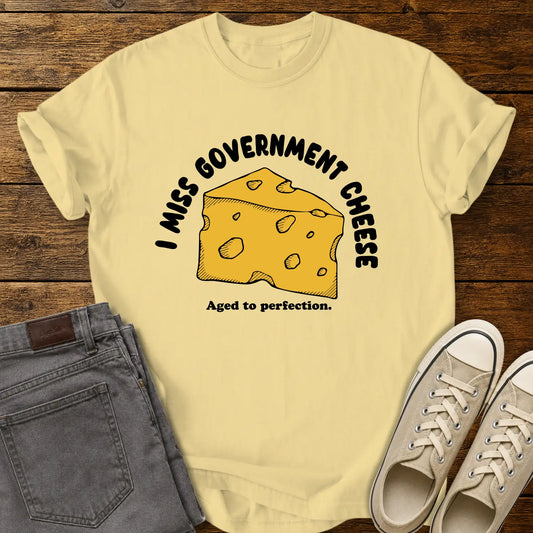 butter yellow T-shirt mockup featuring a hand-drawn wedge of yellow cheese with the phrase “I MISS GOVERNMENT CHEESE” arched above and “Aged to perfection.” below. The shirt is displayed on a rustic wood background alongside folded gray jeans and cream-colored canvas sneakers.