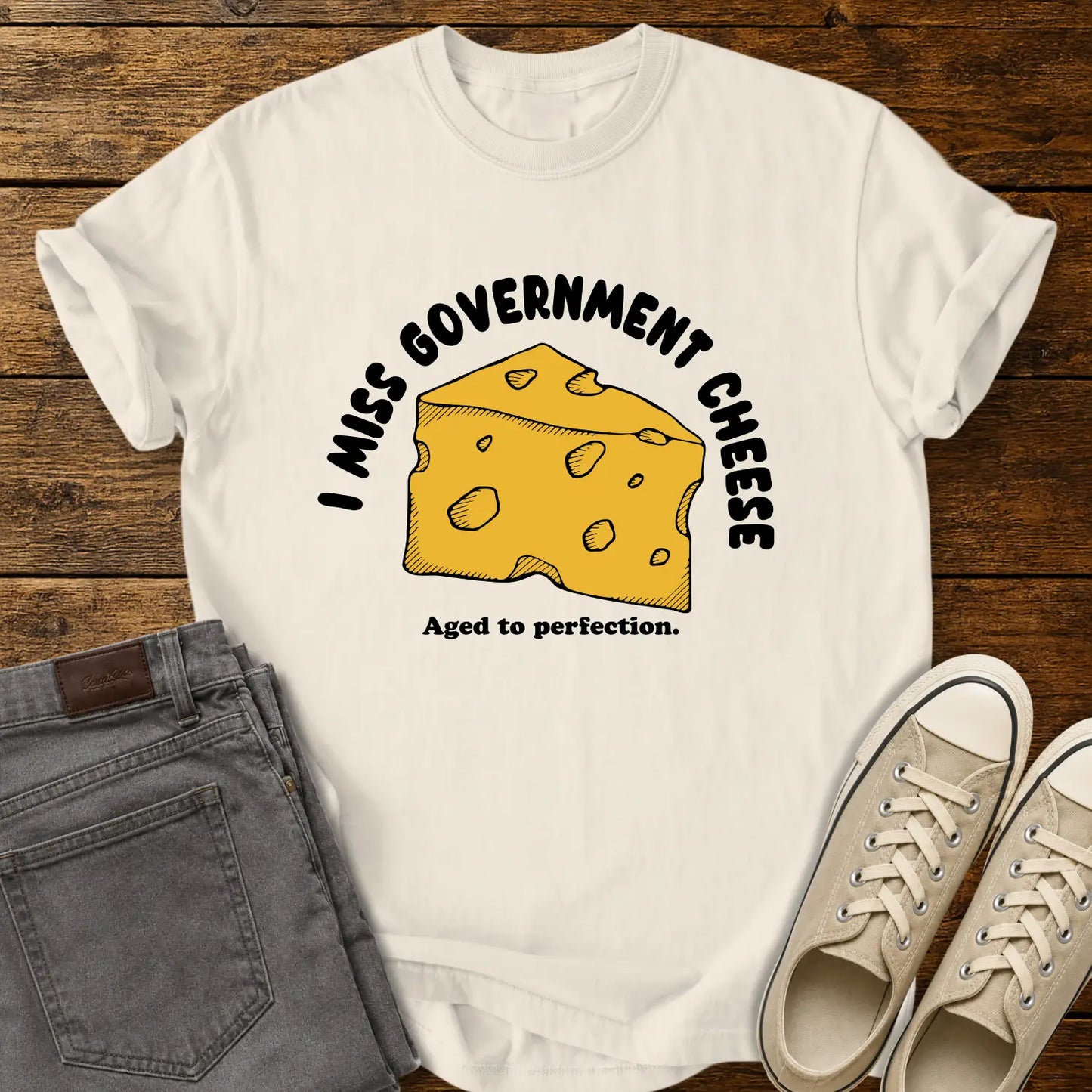 ivory T-shirt mockup featuring a hand-drawn wedge of yellow cheese with the phrase “I MISS GOVERNMENT CHEESE” arched above and “Aged to perfection.” below. The shirt is displayed on a rustic wood background alongside folded gray jeans and cream-colored canvas sneakers.